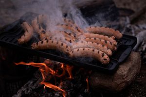 Sausages cooking on a grill plate over open flames with smoke rising in a rustic outdoor setting.