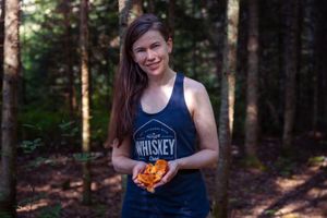 A runner wearing a tank top poses in a forest setting while holding an orange object in their hands.