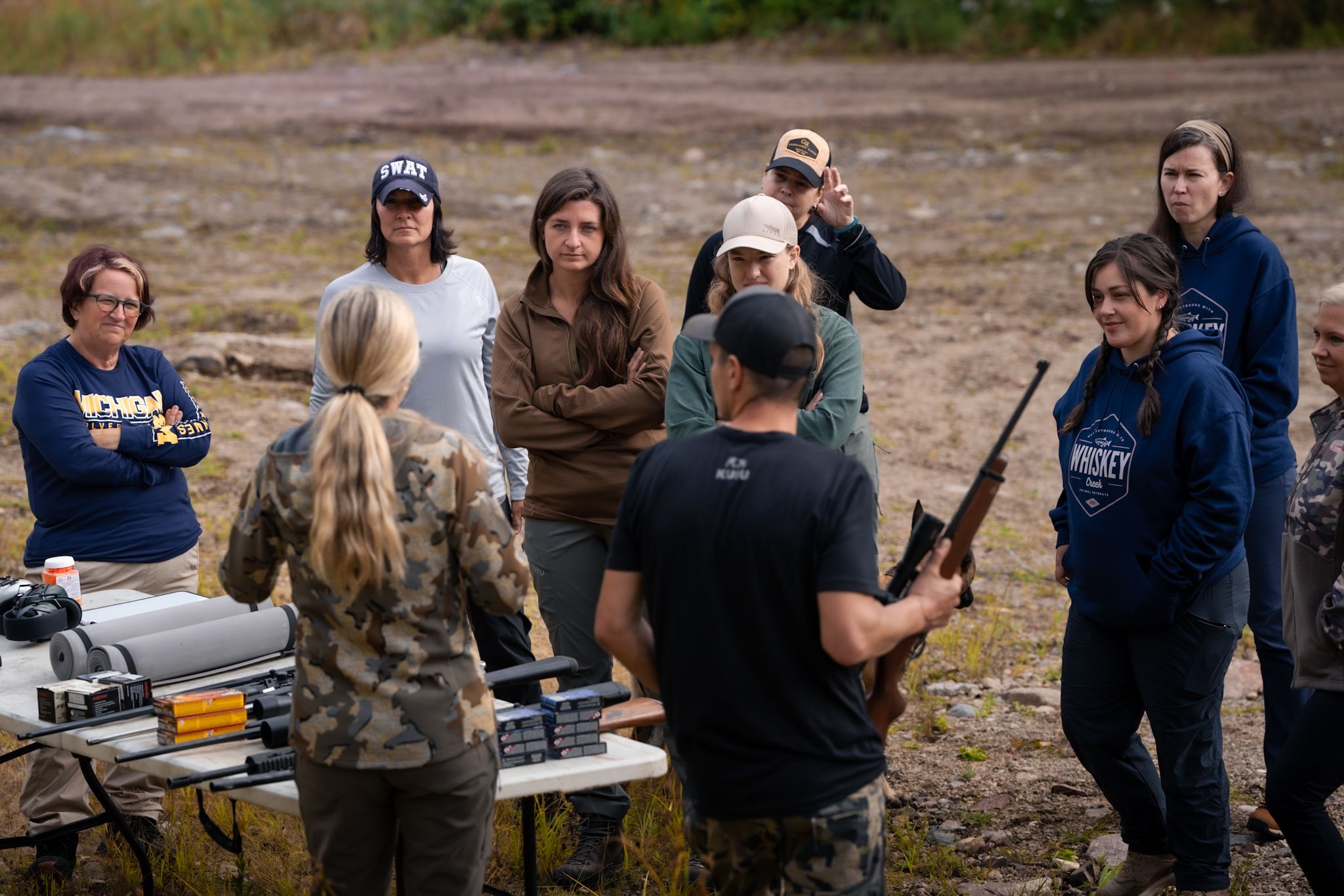 A group gathers around a table outdoors for instruction while holding sporting equipment and wearing casual attire.