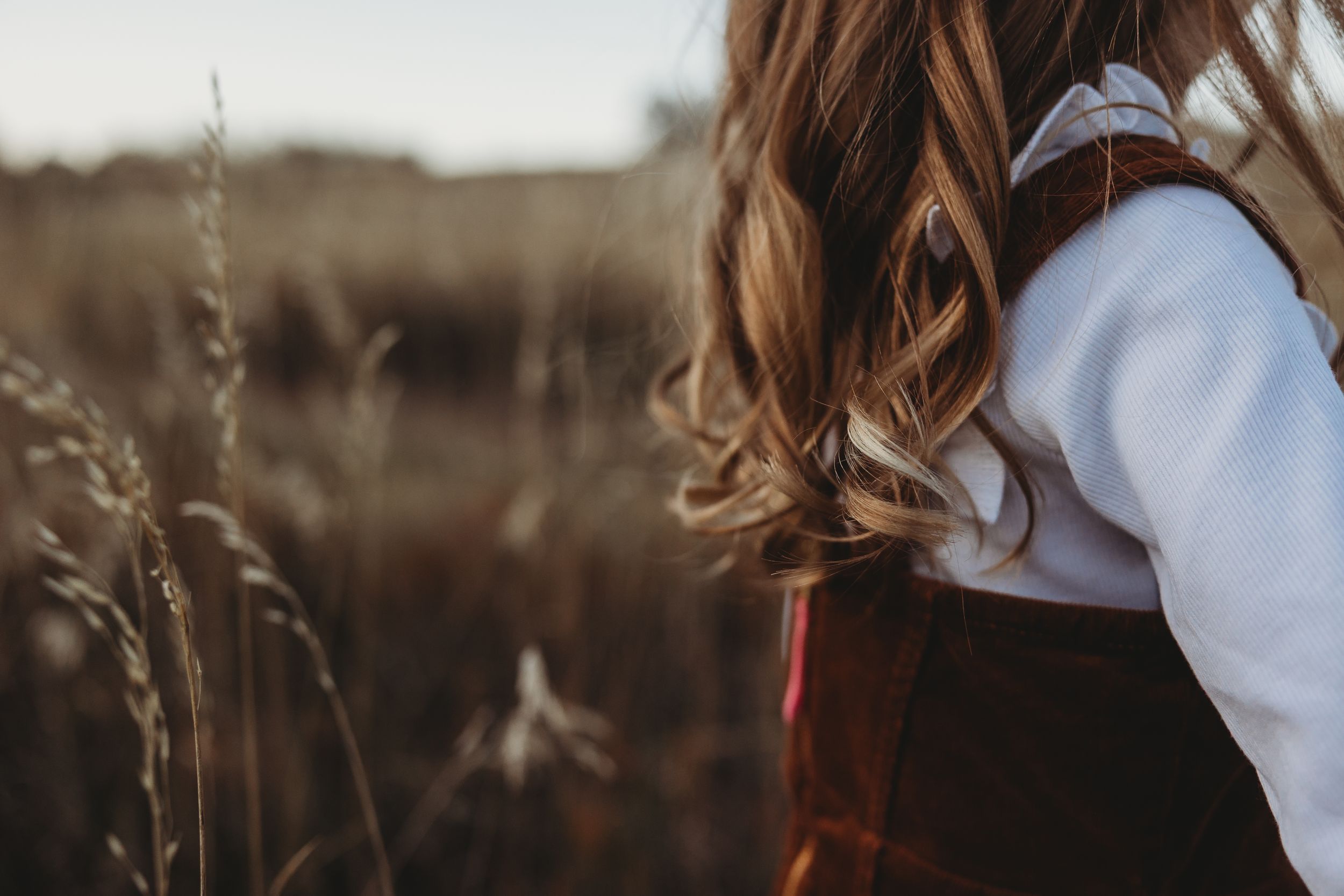 Young woman with long wavy brown hair standing in a golden field wearing a rust jumper.