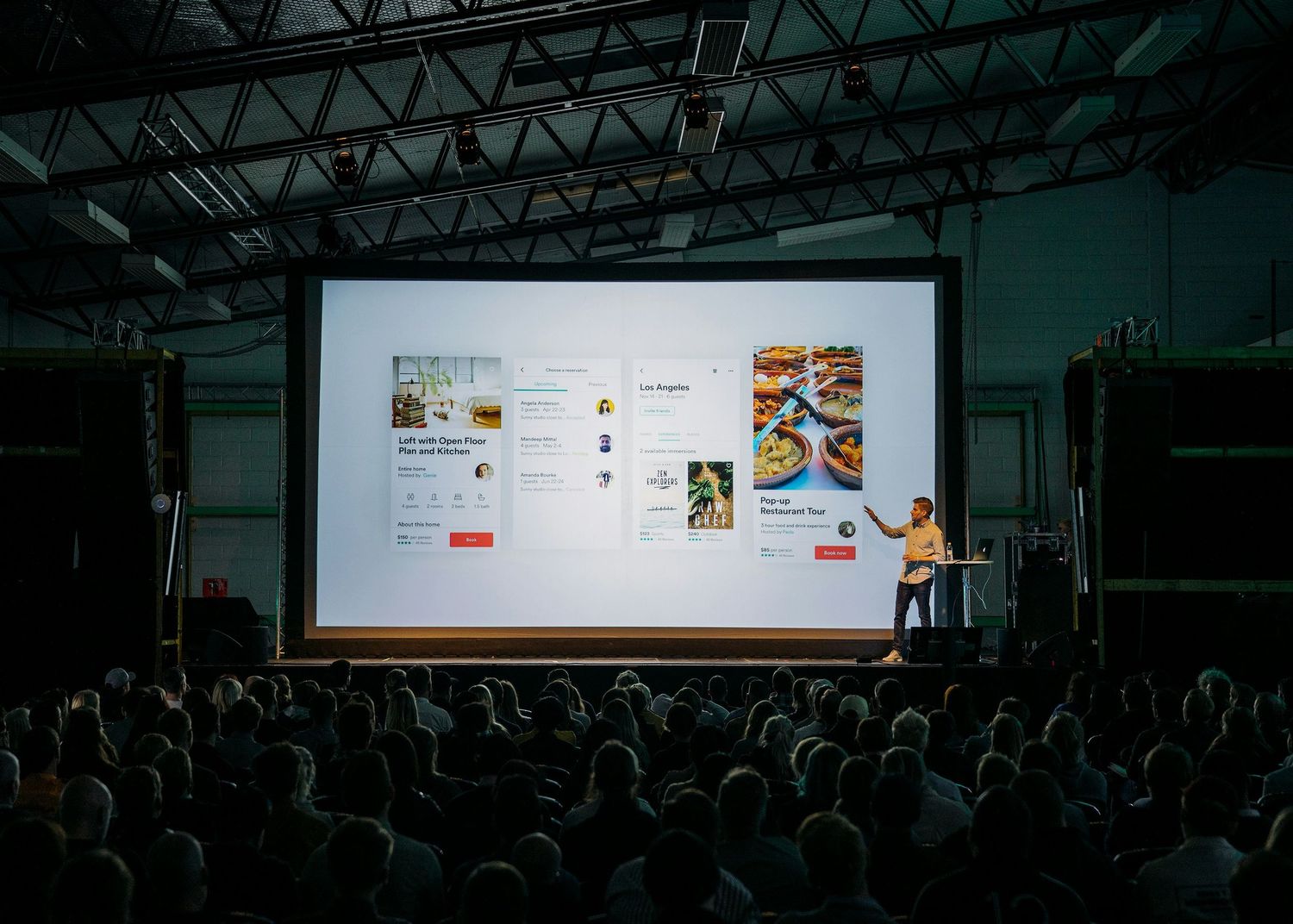 A presenter gives a presentation on a large projection screen to an audience seated in a dark auditorium.