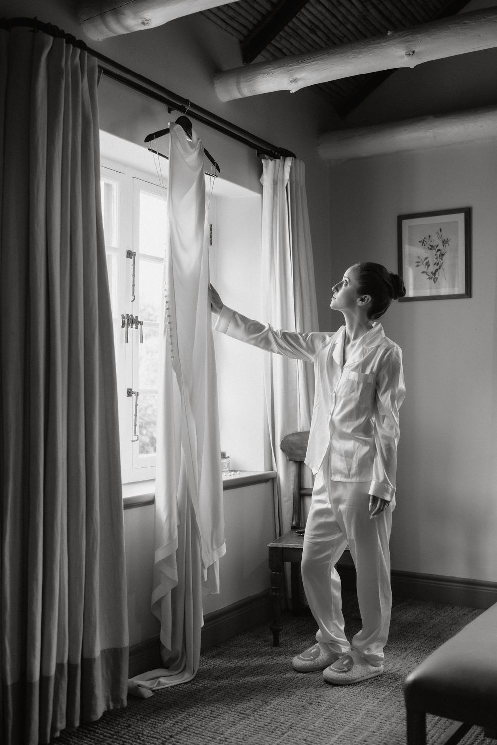 A white wedding dress hangs by a window while someone in white attire inspects it in a room with natural light.