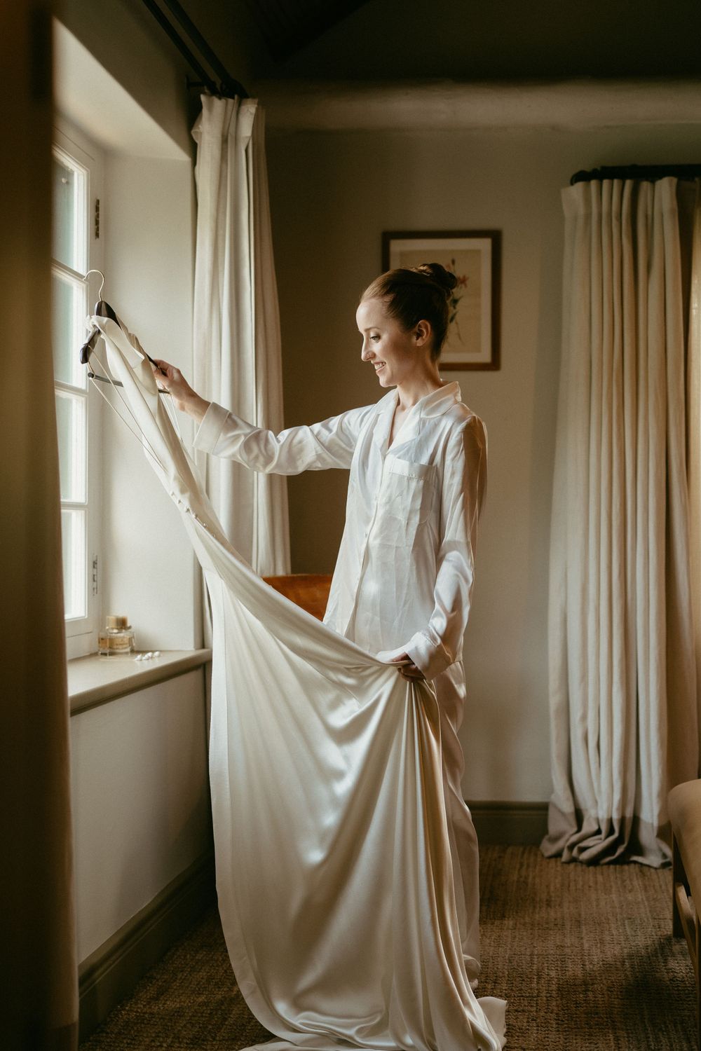 A person in white examines a wedding dress by natural window light in a serene room.