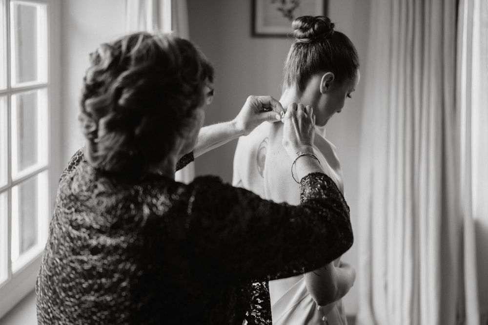 Black and white photo of a bride getting ready with assistance in a sunlit room.