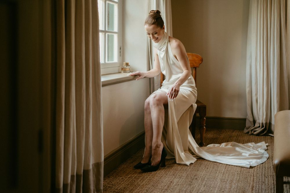 Someone in a flowing white dress sits gracefully by a window while putting on shoes during wedding preparations.
