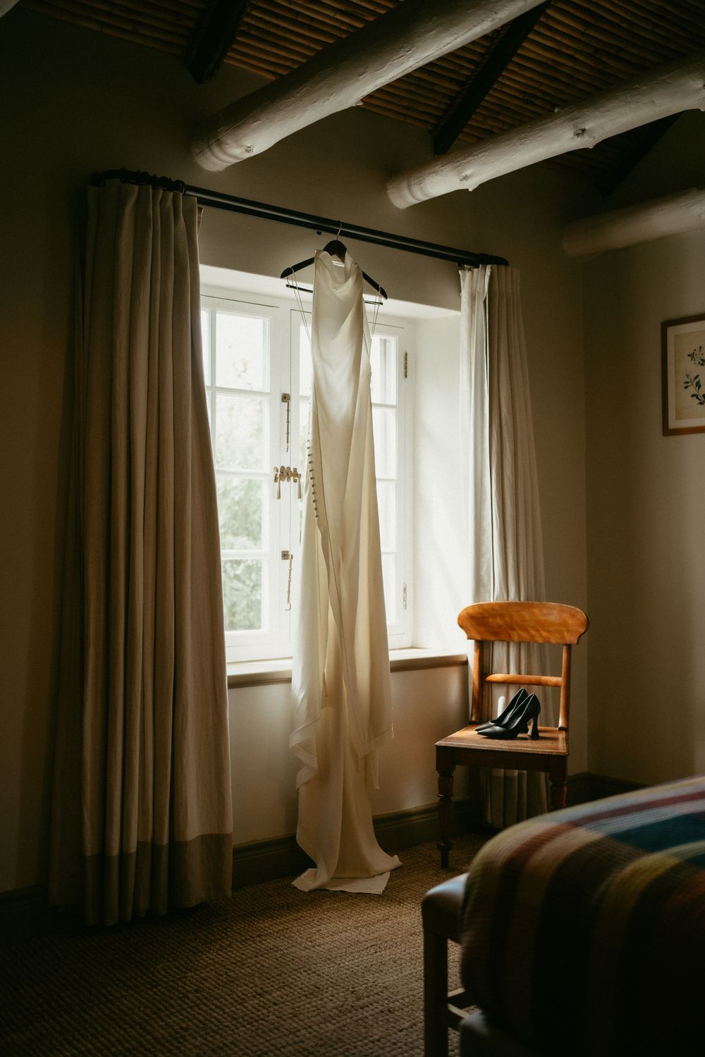 An elegant white wedding dress hangs from a hanger near a window with flowing curtains and exposed wooden beams.