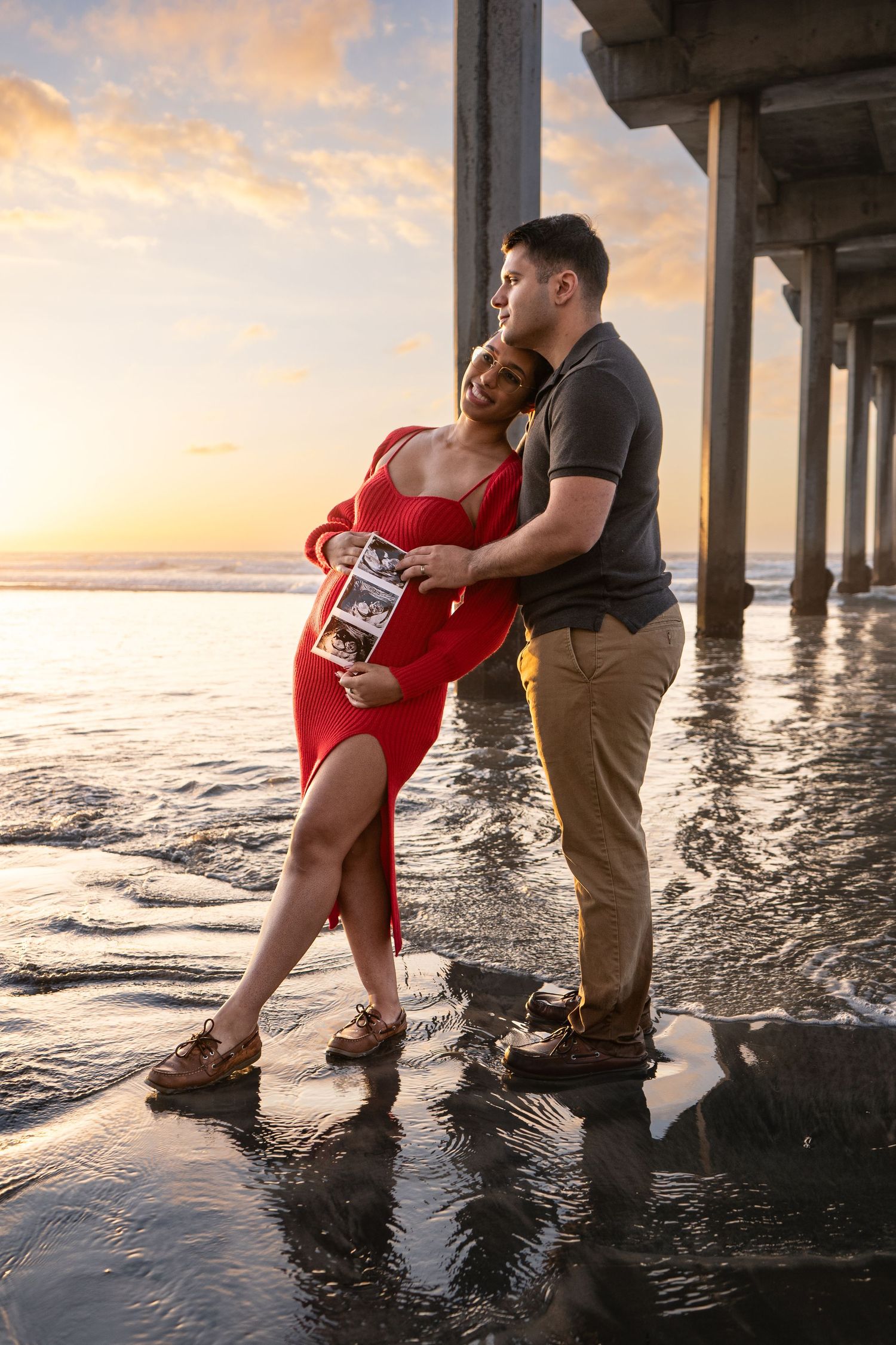 Couple embracing by a pier during sunset with waves lapping at their feet.