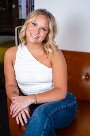 Woman in white one-shoulder top and jeans sits casually on brown leather couch.