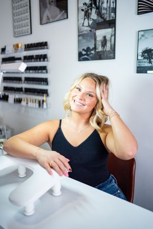 Woman in black tank top at beauty salon desk with nail supplies on wall.