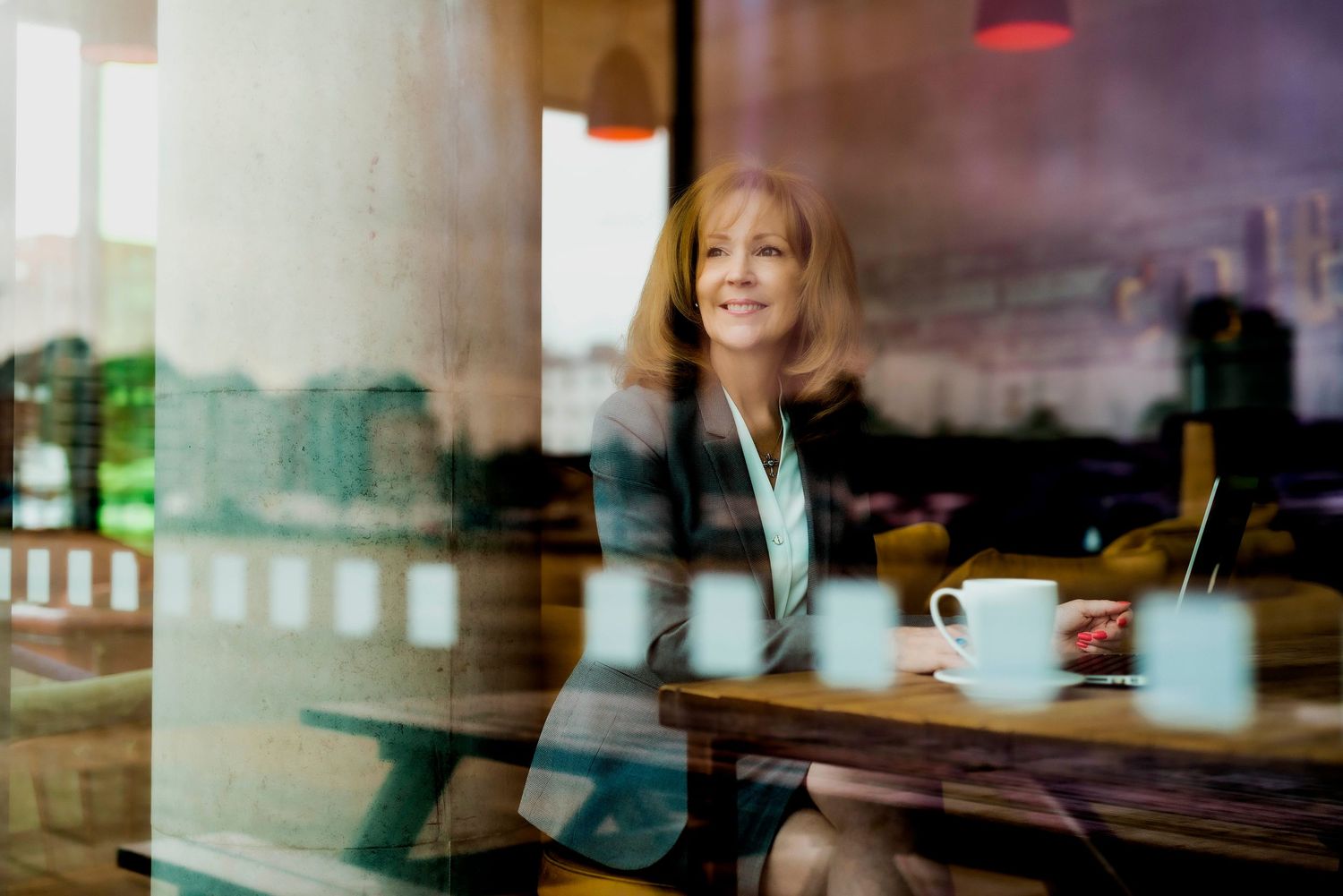 A Businesswoman sits at a cafe table with a coffee cup, viewed through a window with city reflections in the glass.