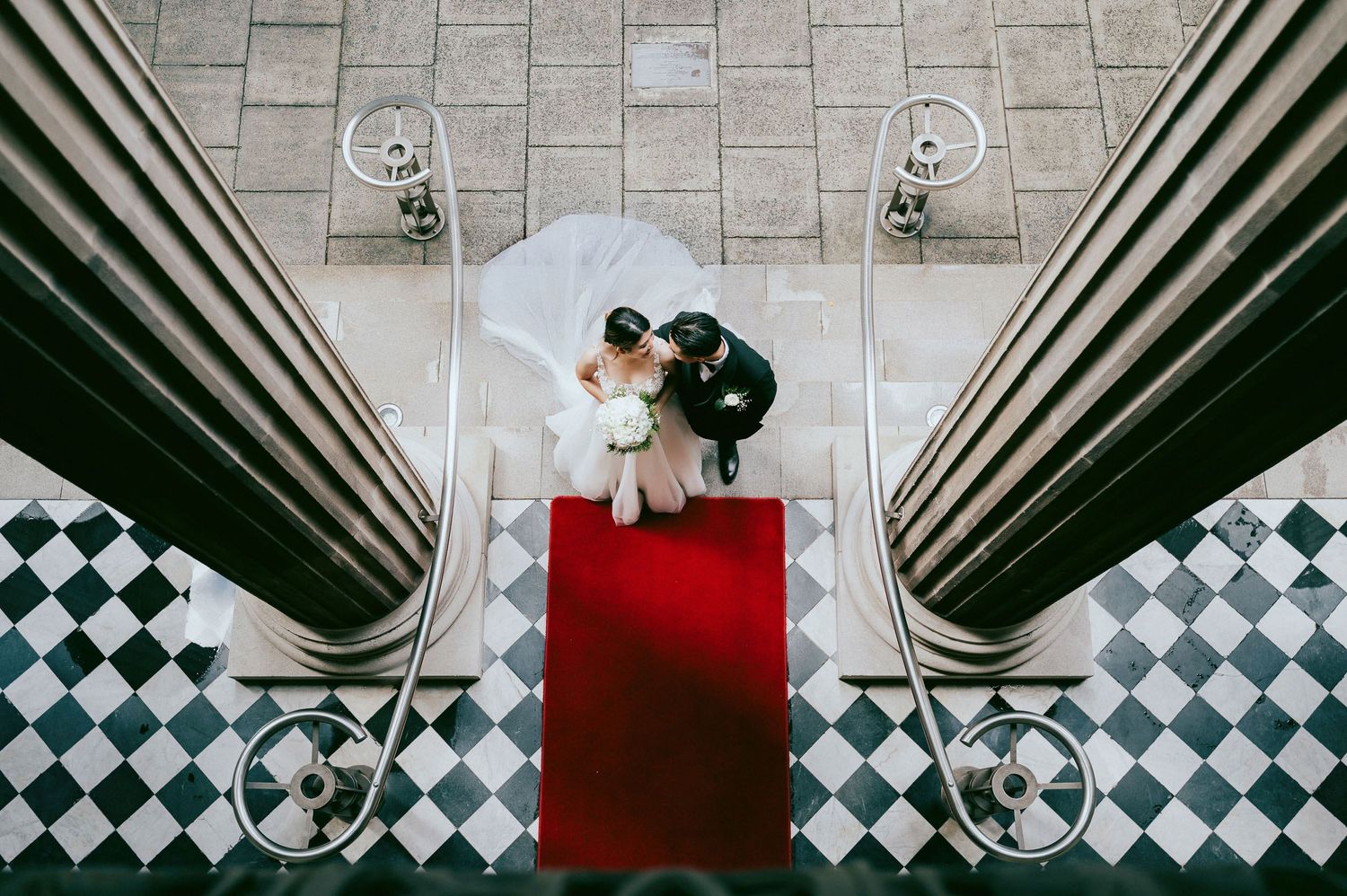 Overhead view of couple on red carpet between classical columns with checkerboard floor.