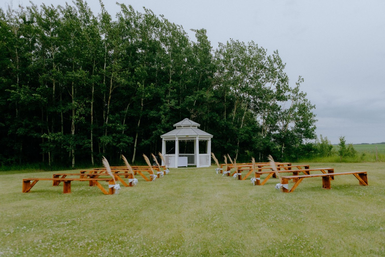 A white gazebo with wooden benches arranged in a semicircle for an outdoor wedding ceremony.