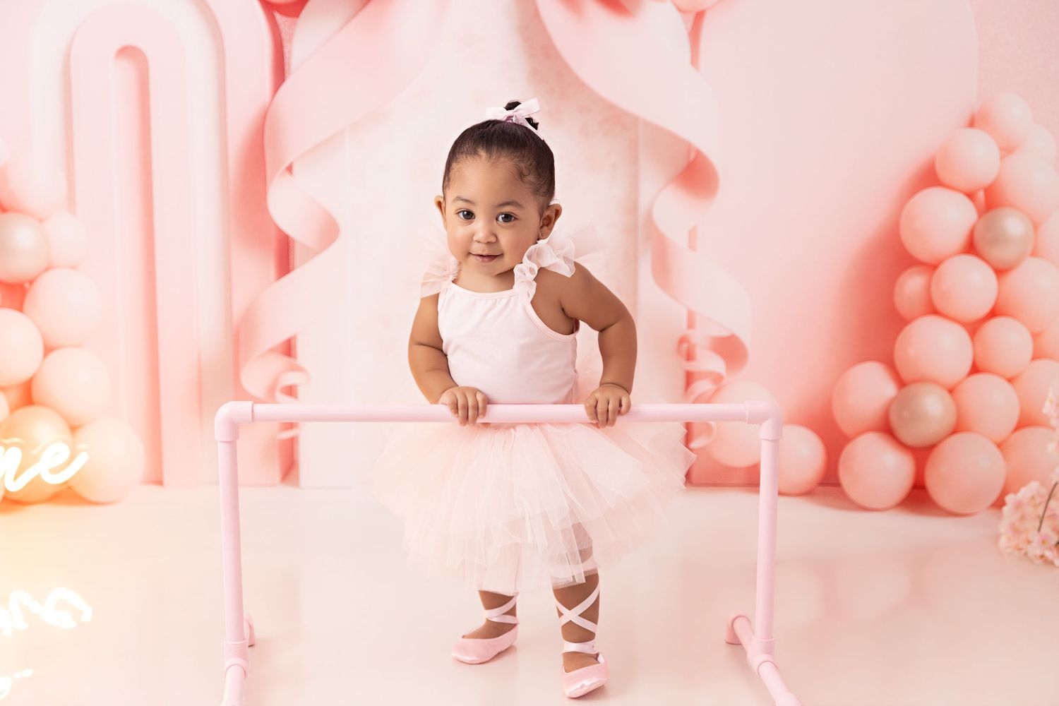 Baby in pink dress sits with cake against backdrop of pink balloons arranged in fan shape for first birthday photoshoot.