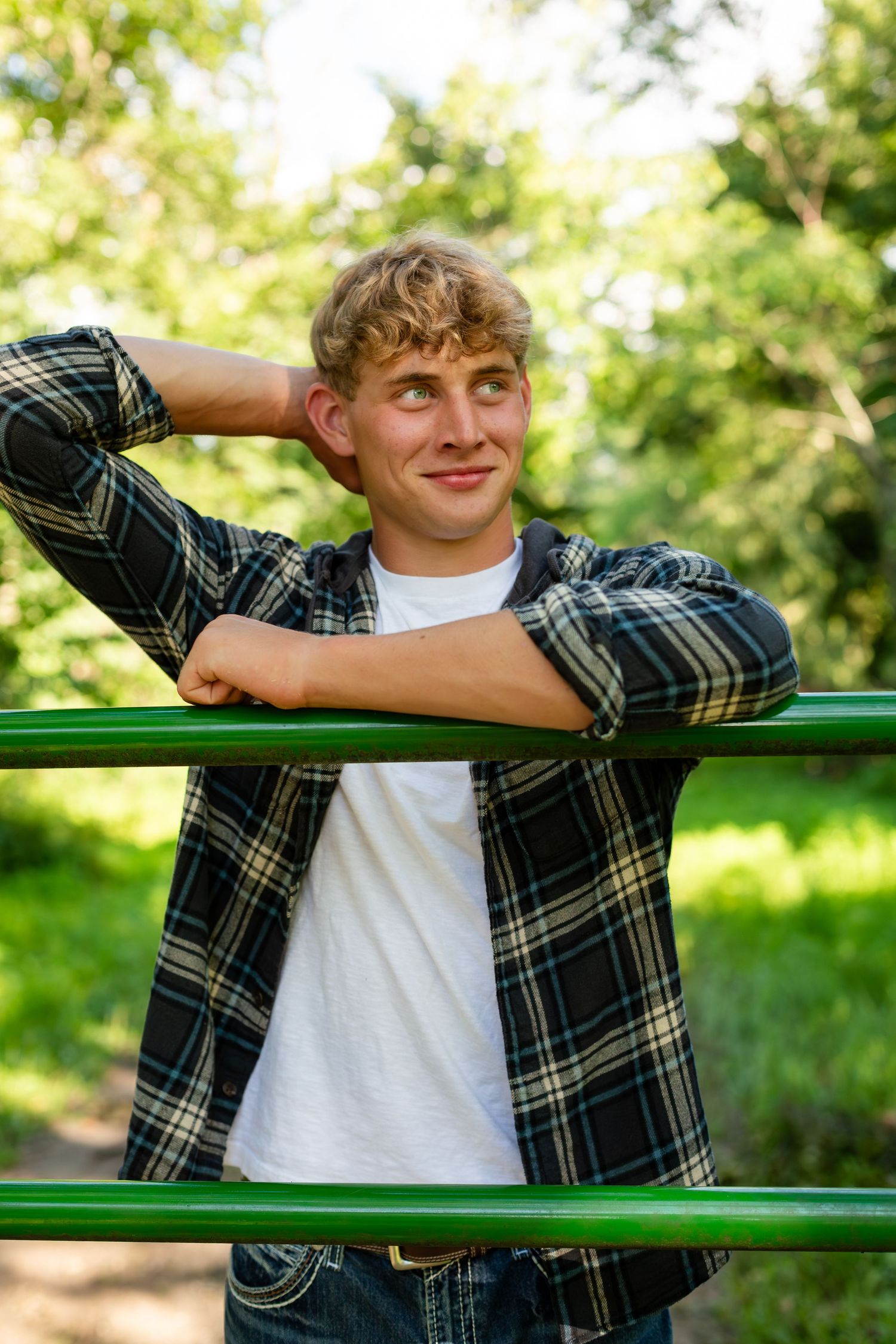 A person relaxes on a green park bench while wearing a plaid flannel shirt and white t-shirt on a sunny day.