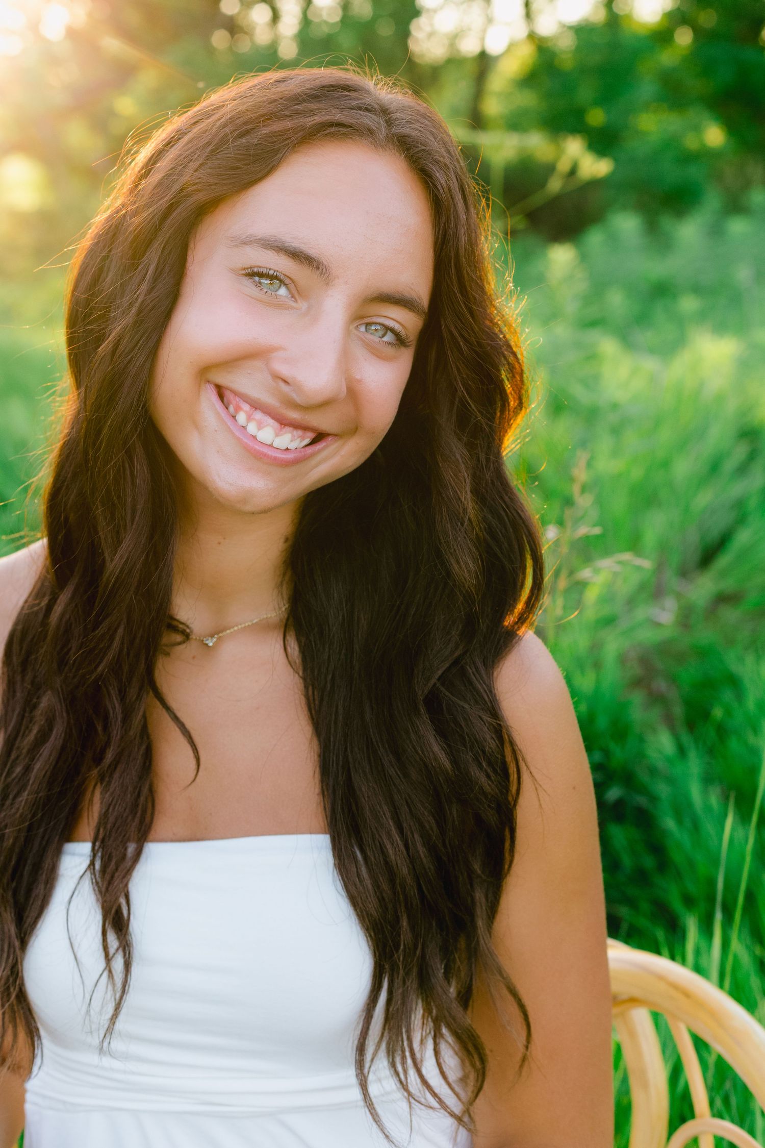 Portrait of a smiling person with long dark hair wearing a white top in a sunlit outdoor setting with greenery.