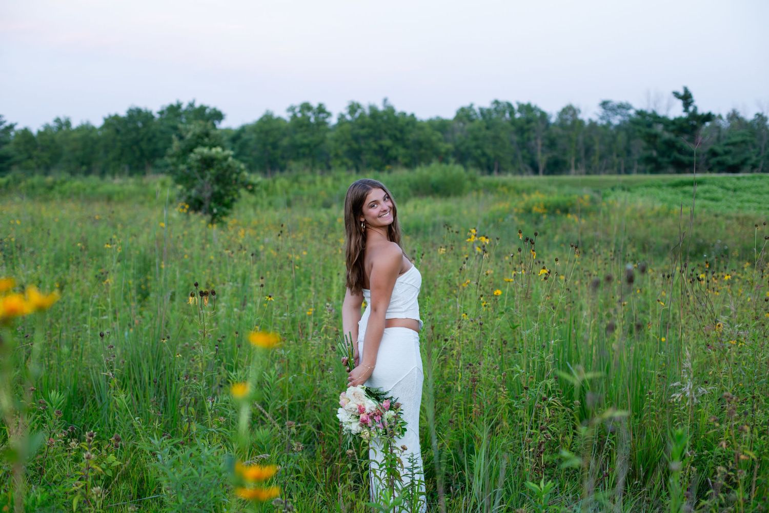 Woman in white dress walking through a meadow of wildflowers and tall grass on a bright summer day.