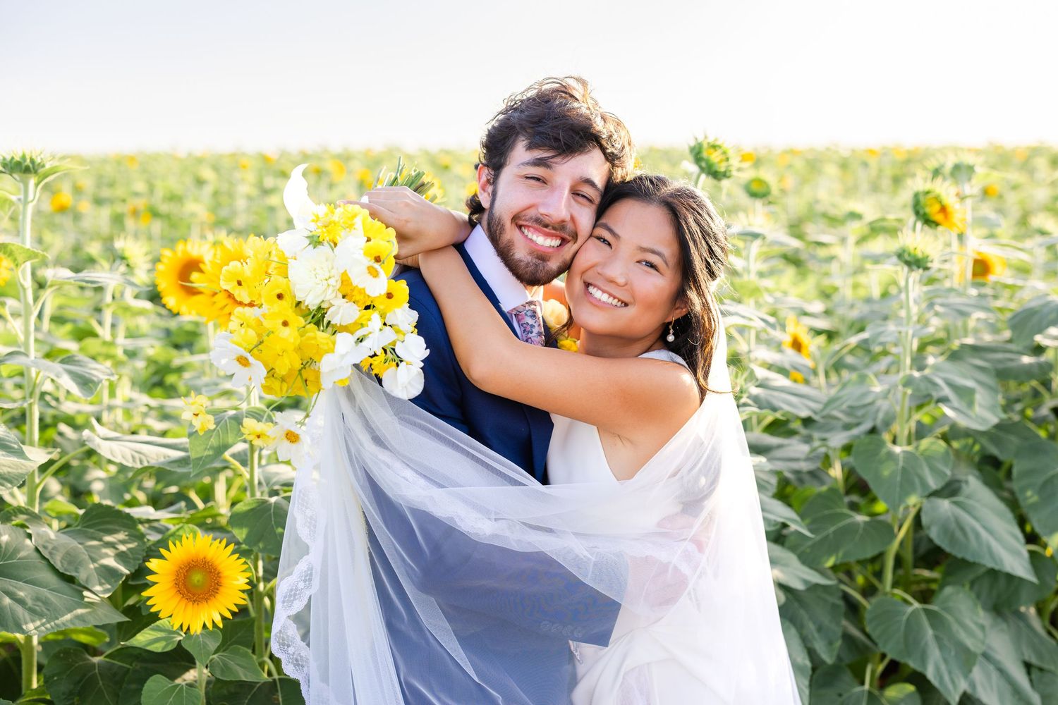 Happy couple embraces while laughing together in a vibrant field of tall blooming sunflowers on a sunny day.