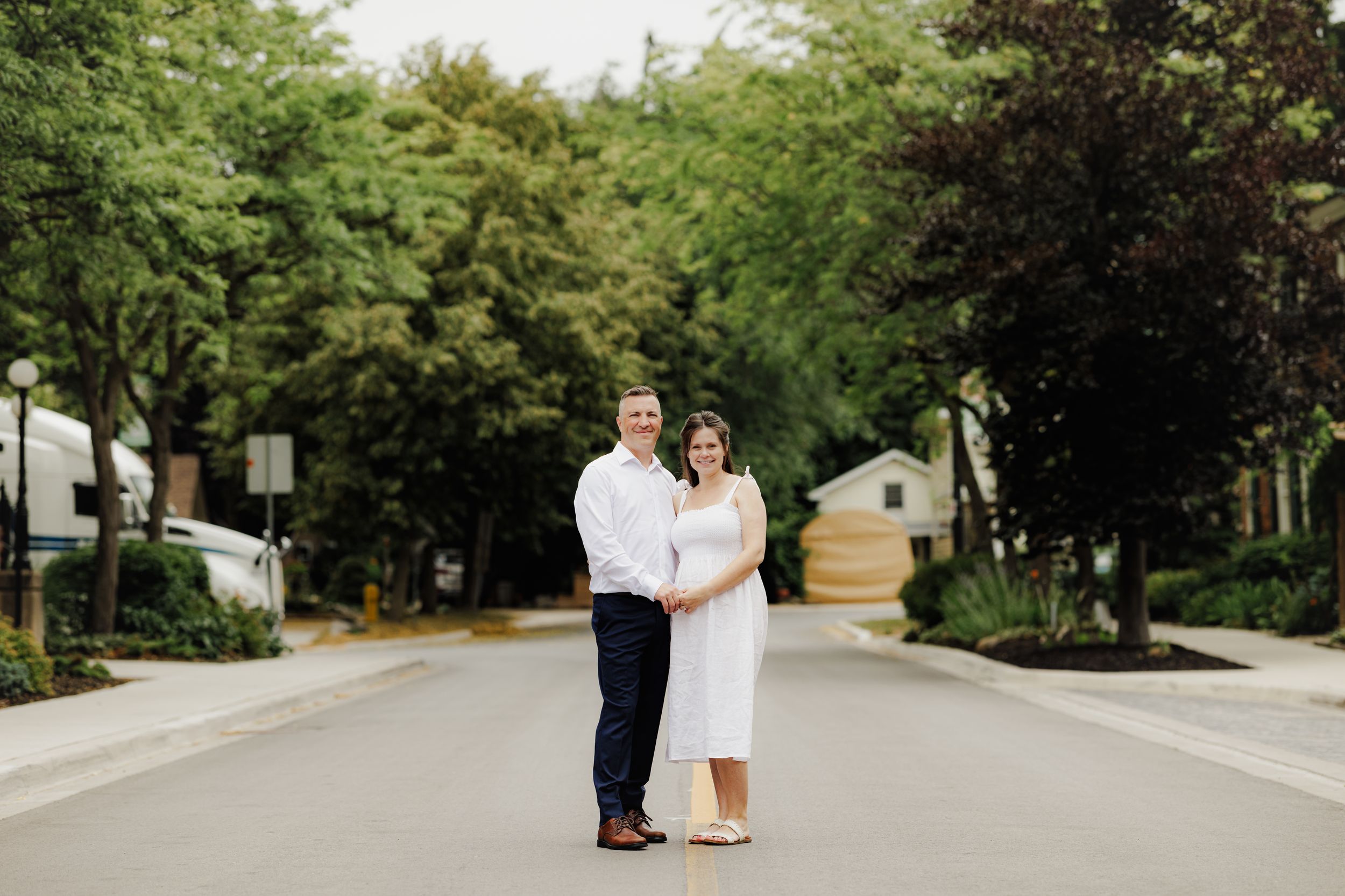 A couple in wedding attire stands together on a tree-lined residential street in summer.