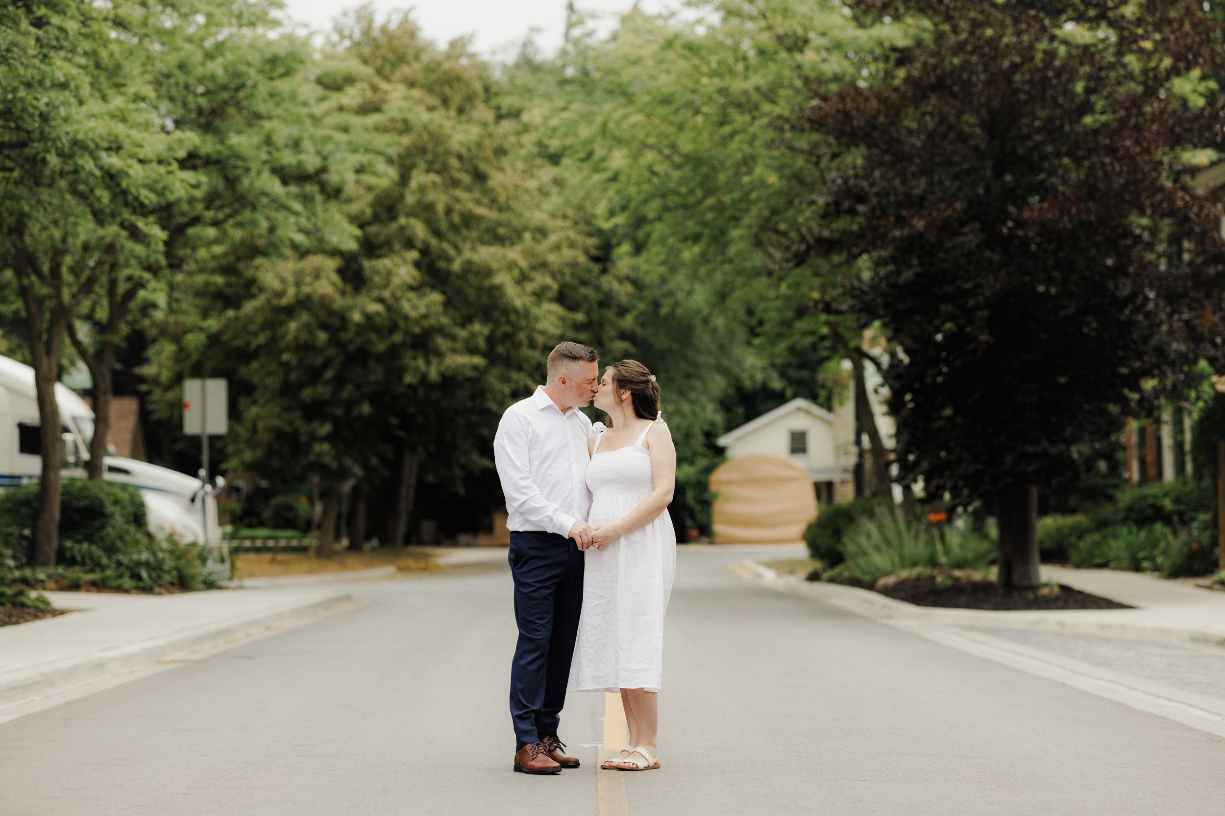 Couple in wedding attire share an intimate moment while walking on a tree-lined path in a park setting.