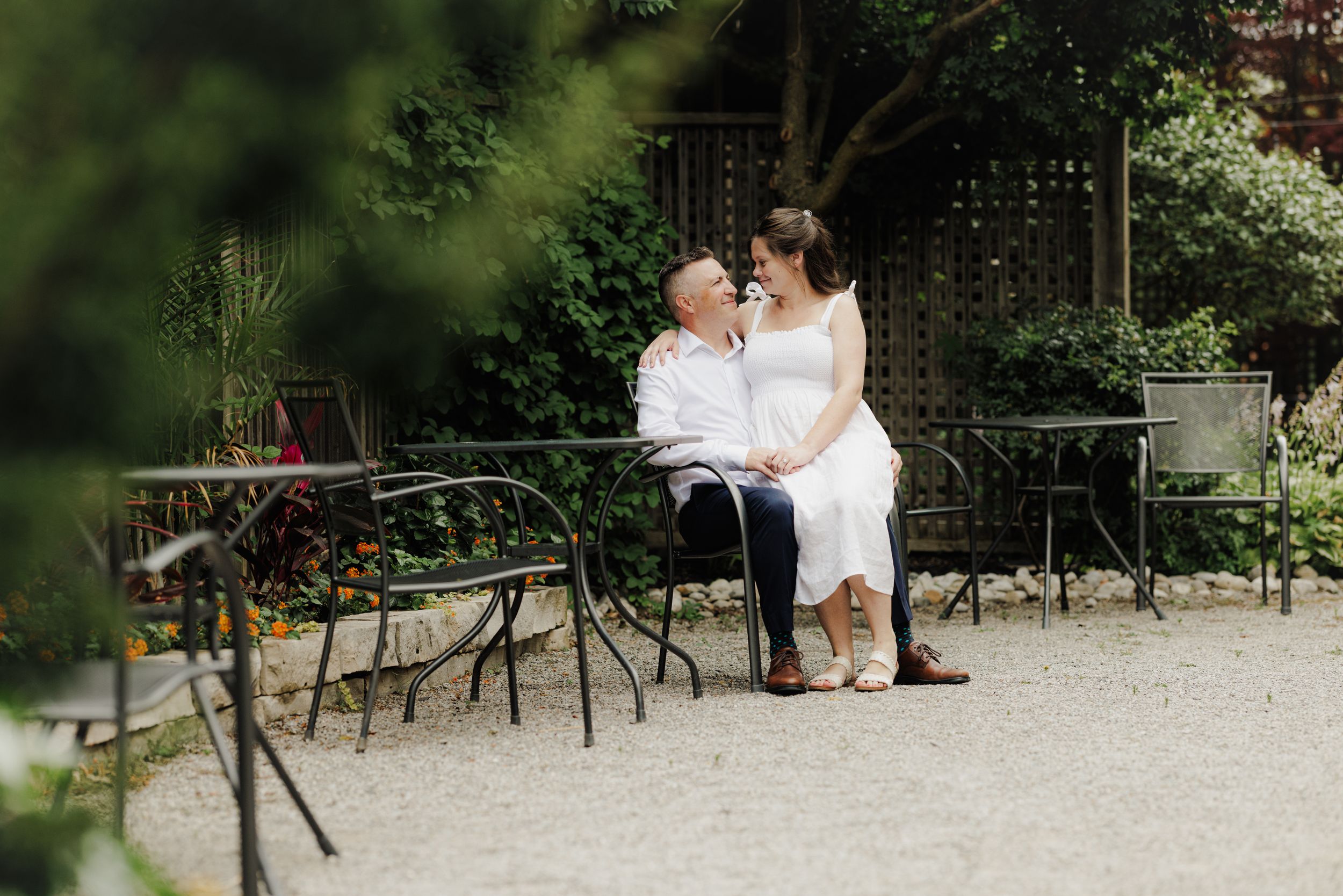 Two people sit together on a wrought iron bench in a serene garden setting with lush greenery and gravel paths.