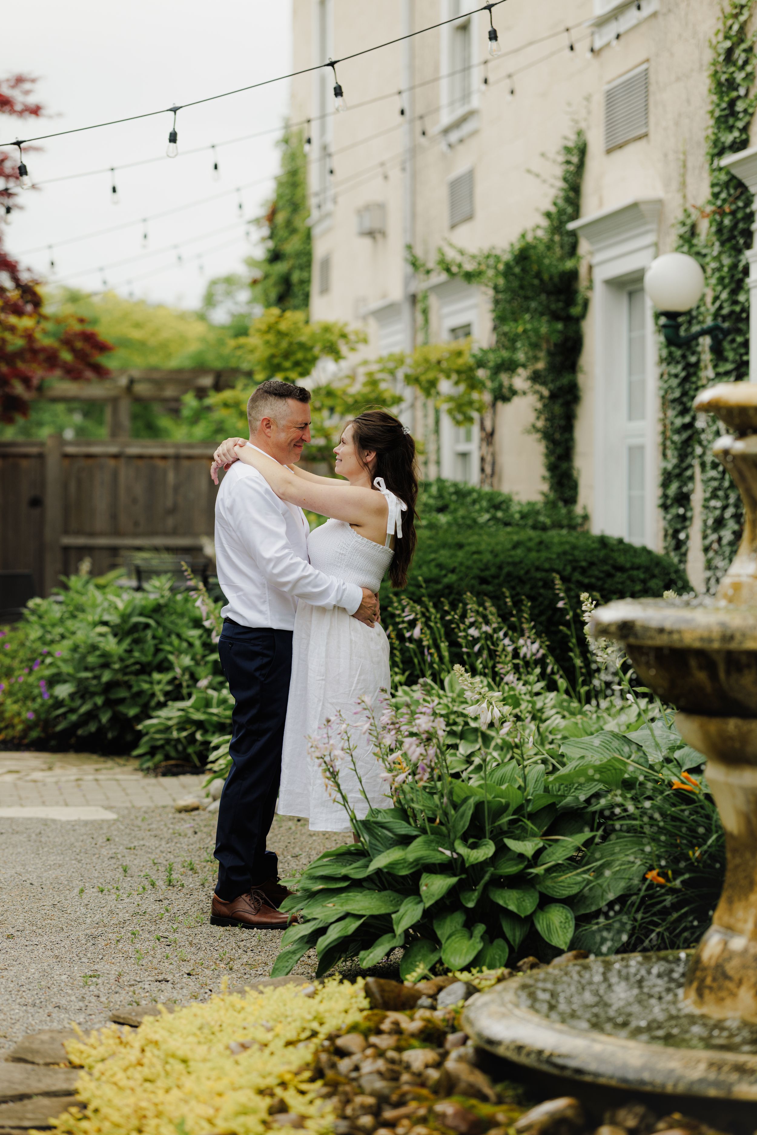 A couple shares a romantic moment in a garden setting with string lights and a stone fountain near a white building.