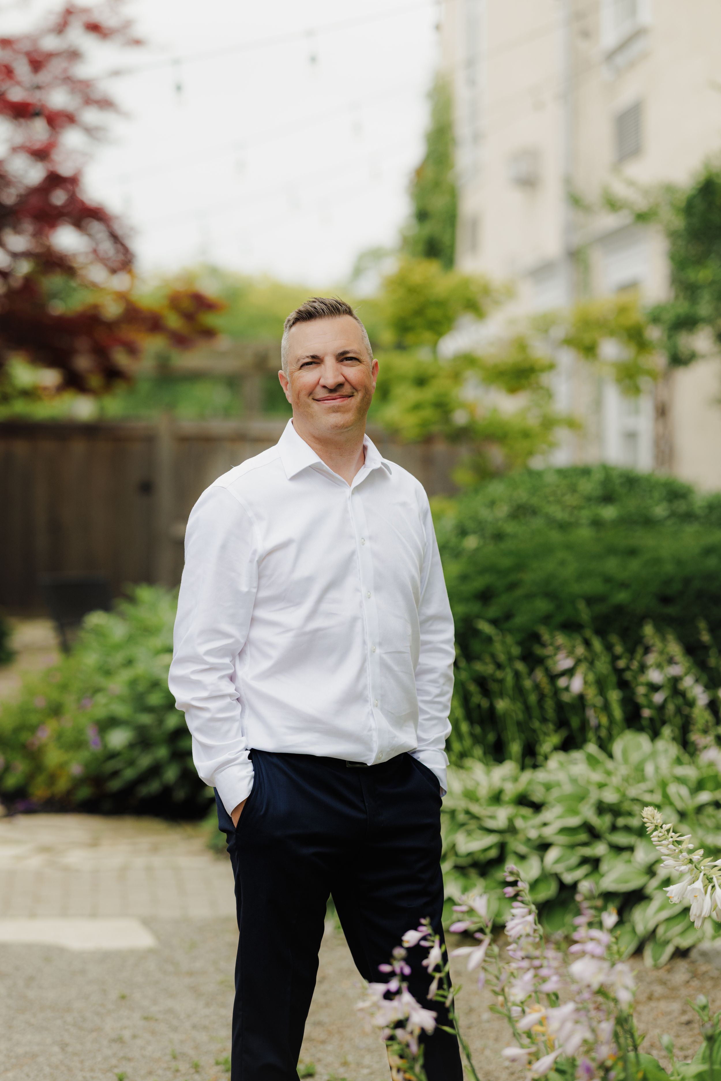 Someone dressed in a white dress shirt and black pants stands in a garden setting with lush greenery in the background.