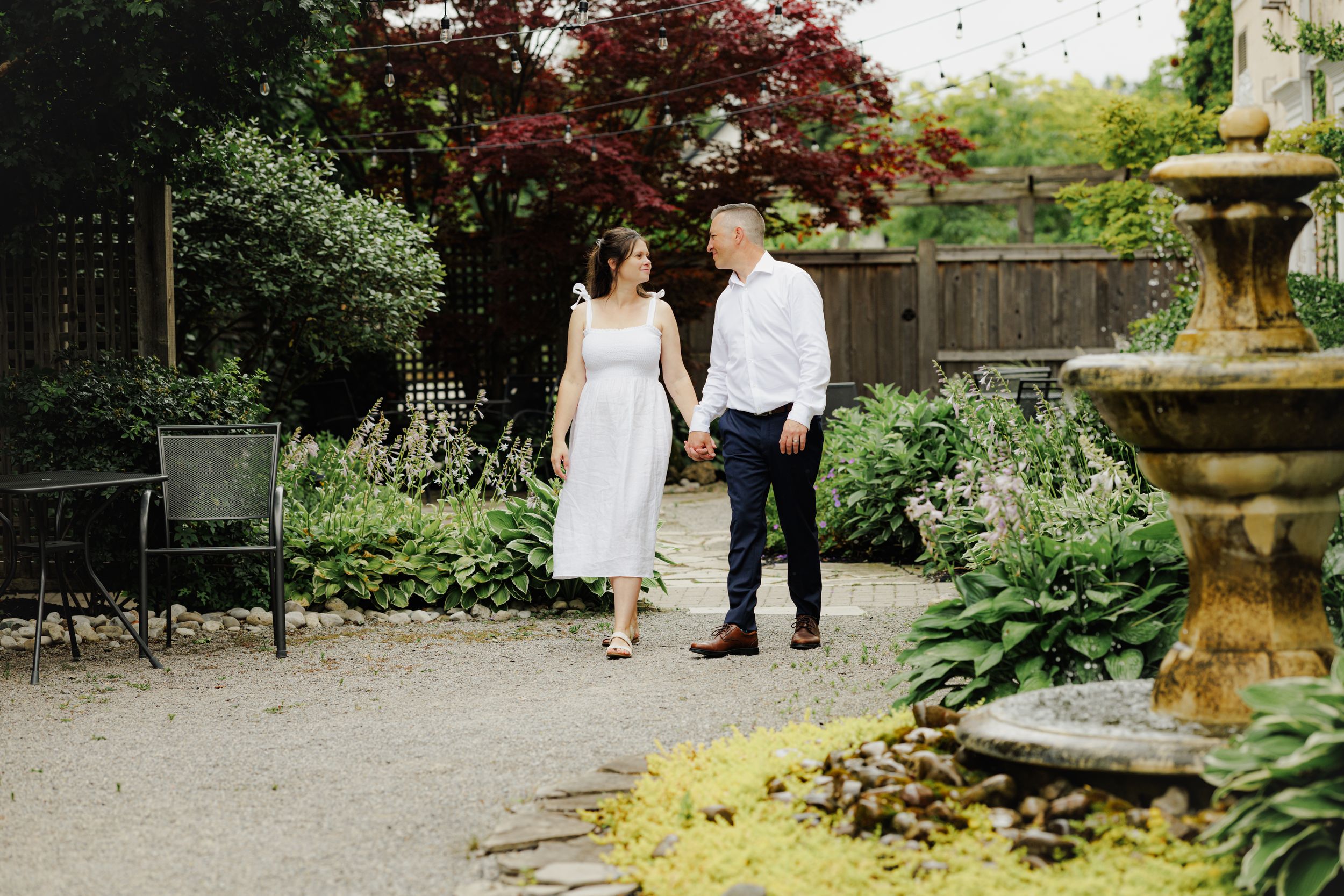 A couple walks together in a garden with a stone fountain, lush greenery and Japanese maple trees.