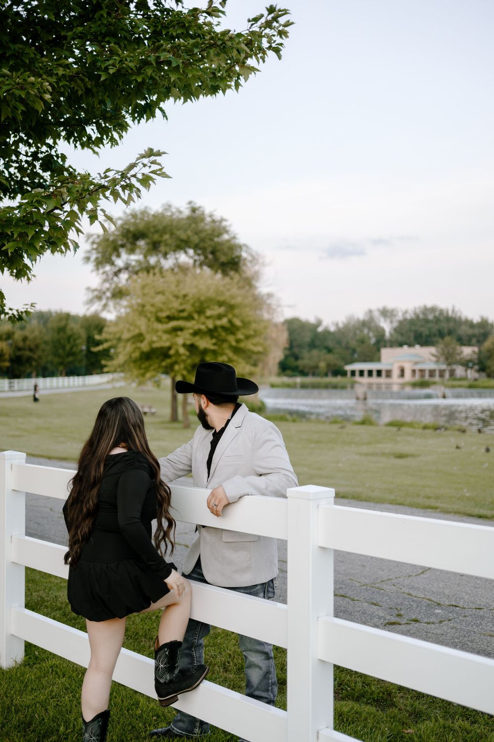 Two people lean against a white fence at a rustic farm property with green grass and trees in background.