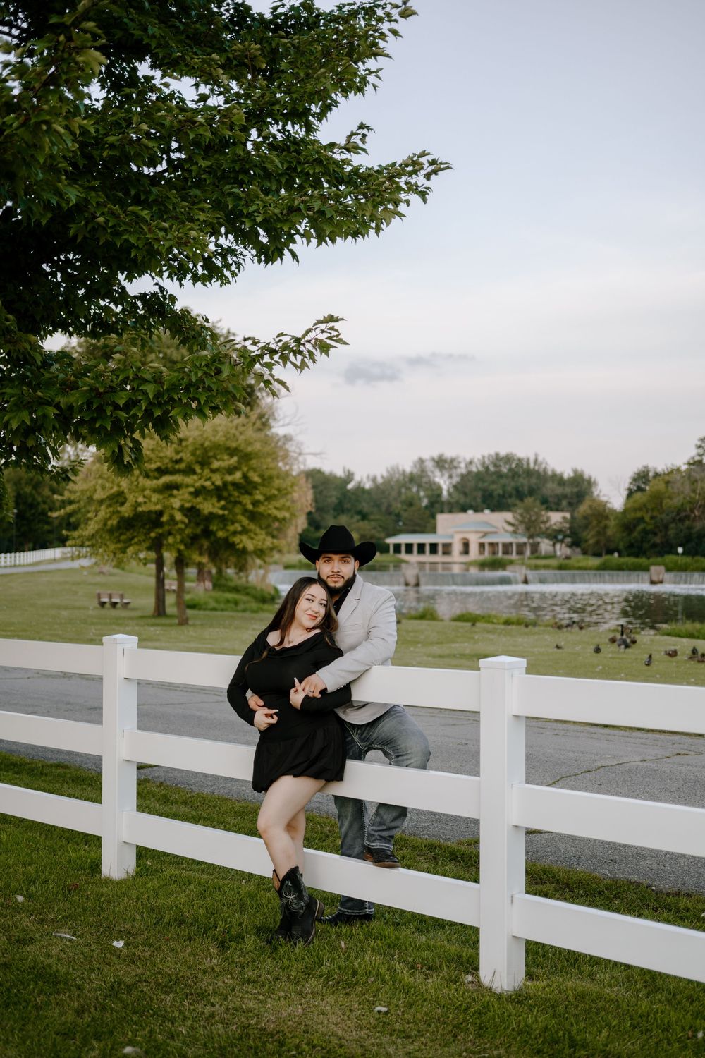 Happy couple sits together and embraces on a rustic white fence in front of trees during an outdoor photo session.