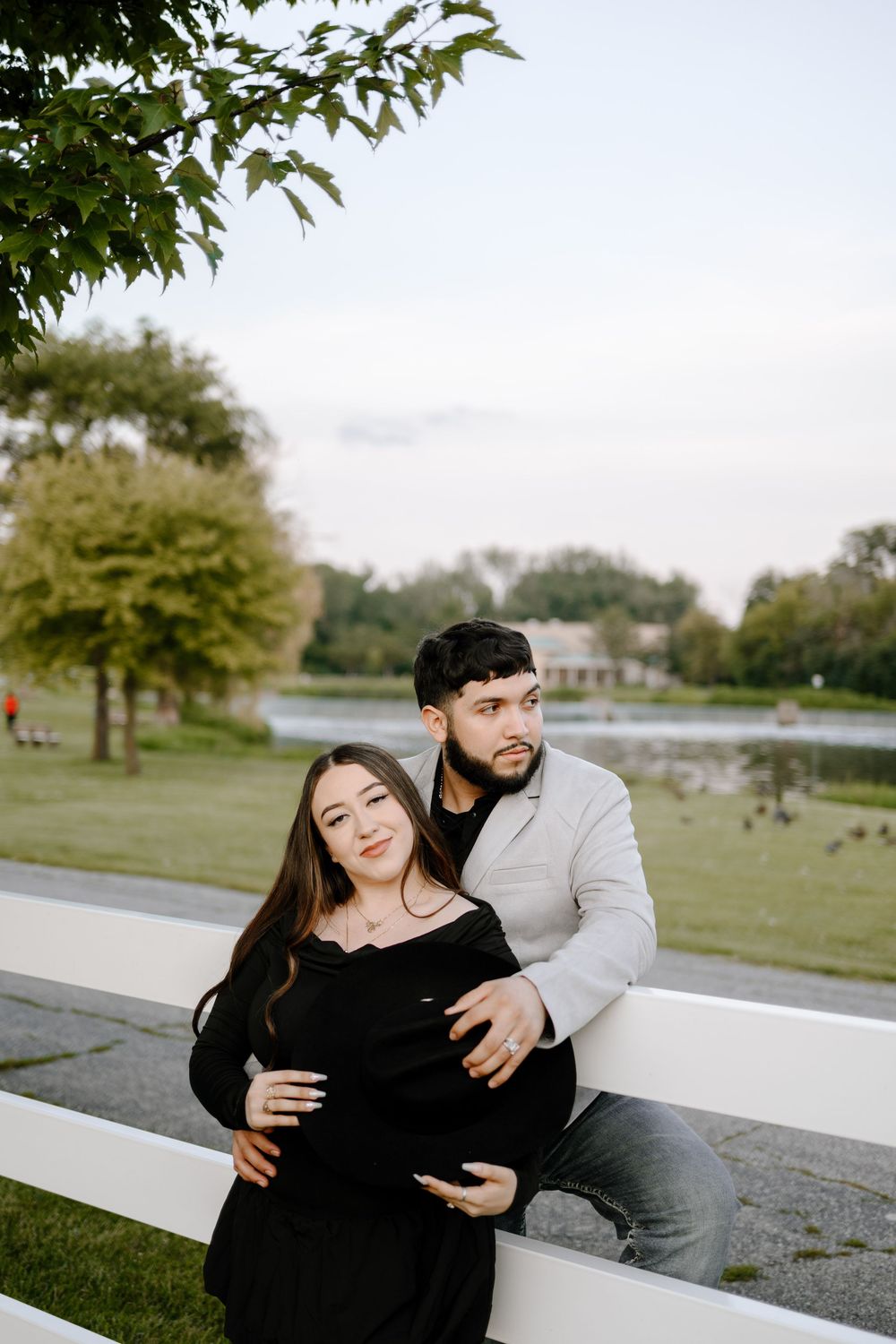 A couple shares an intimate maternity moment together while sitting on a white fence in a park setting.