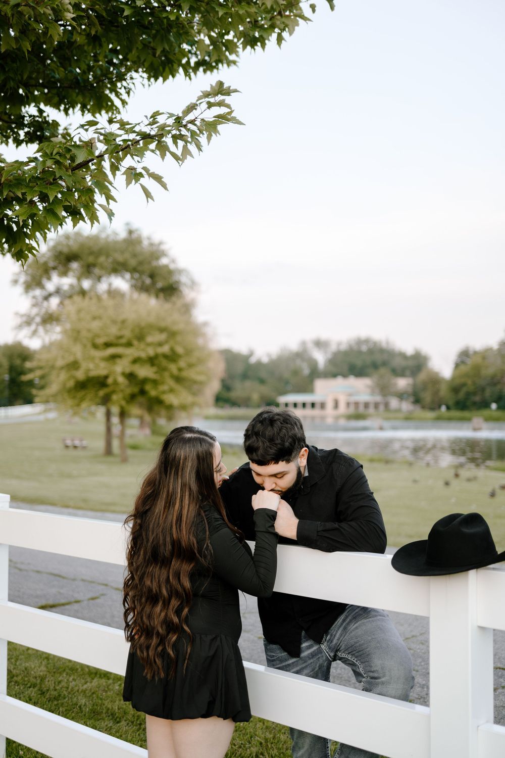 A romantic couple shares a tender moment at a white fence overlooking a scenic lake and park setting.