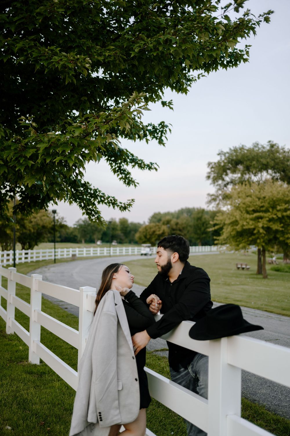 A romantic couple shares tender moments along a white fence on a grassy countryside property.