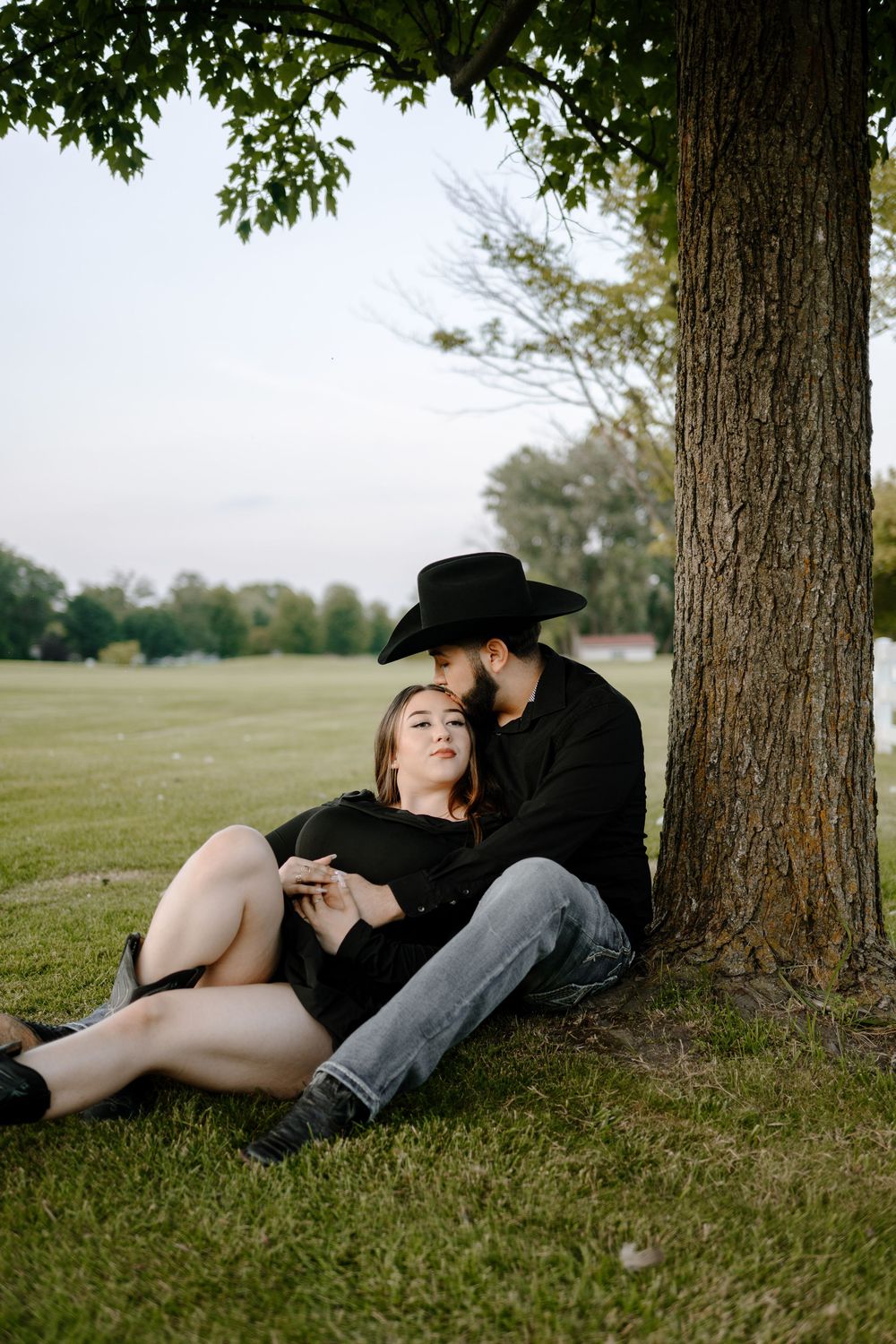 A romantic couple rests under a large tree in a serene park setting during a golden afternoon.
