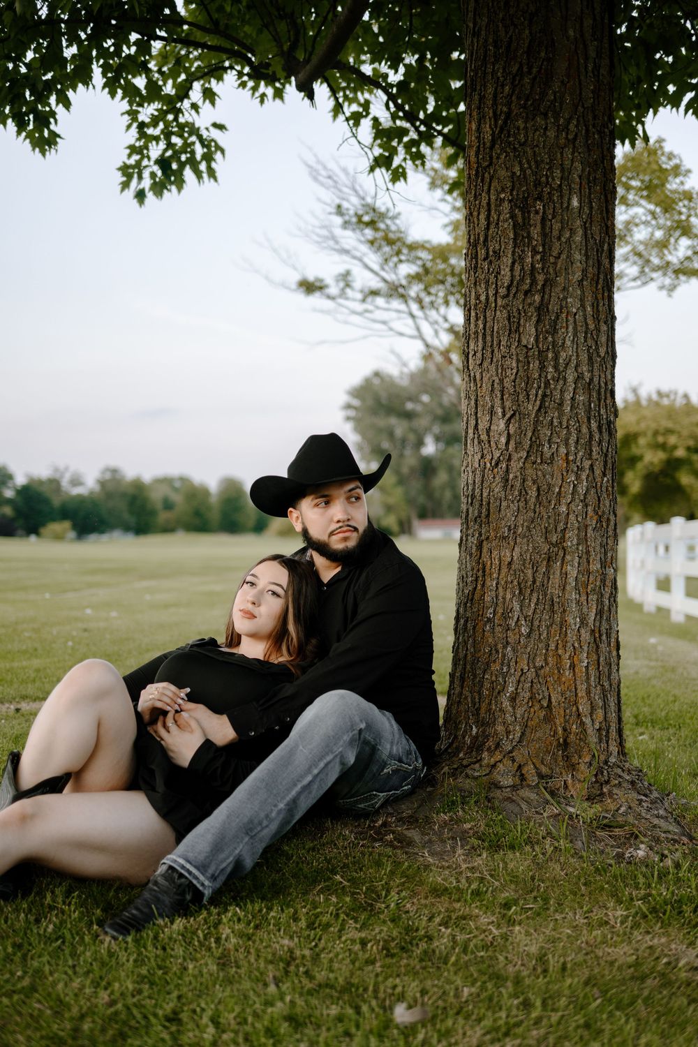 A couple relaxes together under a large tree in a pastoral setting with white fencing visible in the background.