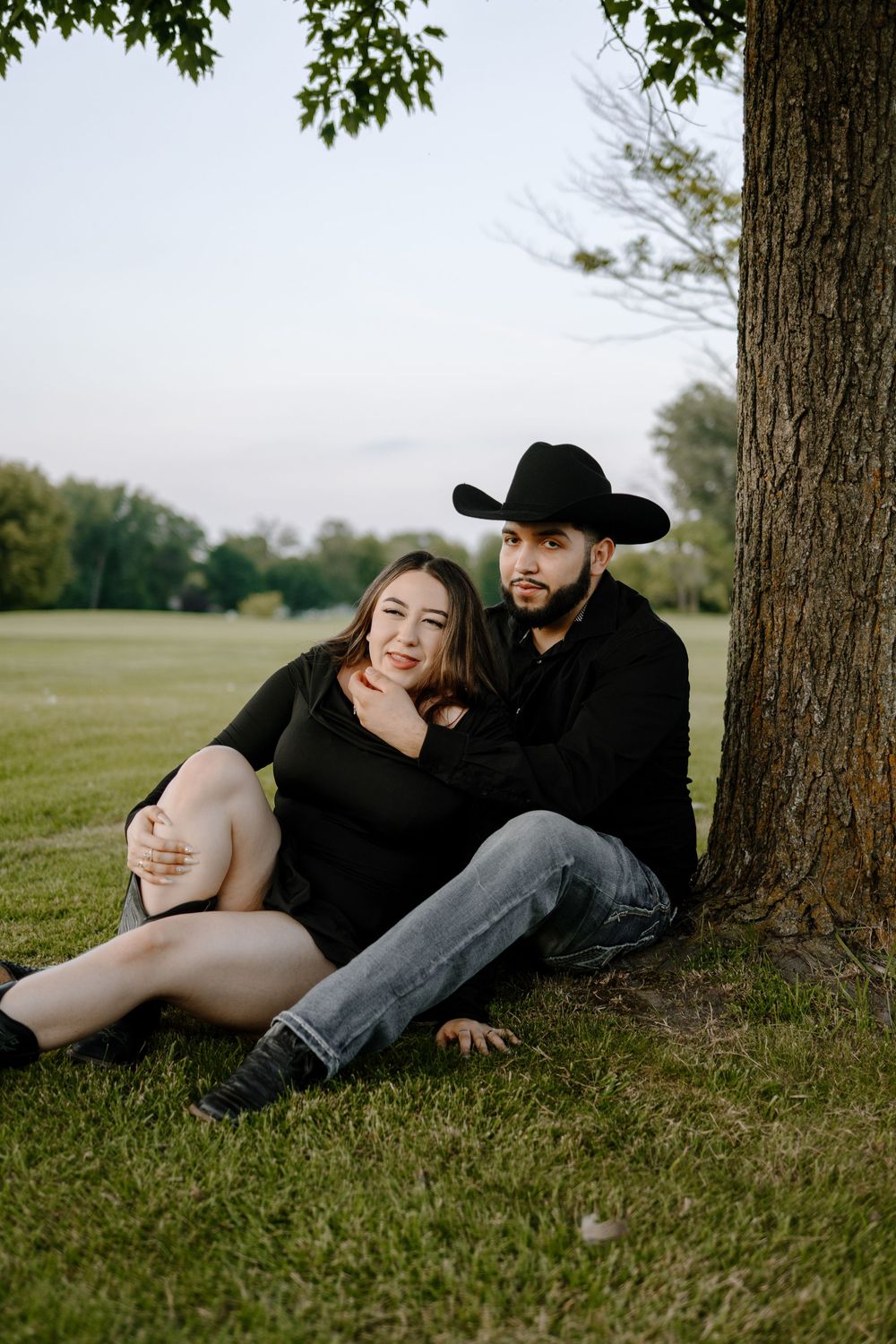 A couple sits together on grass under a tree during sunset, with the man wearing a black cowboy hat.