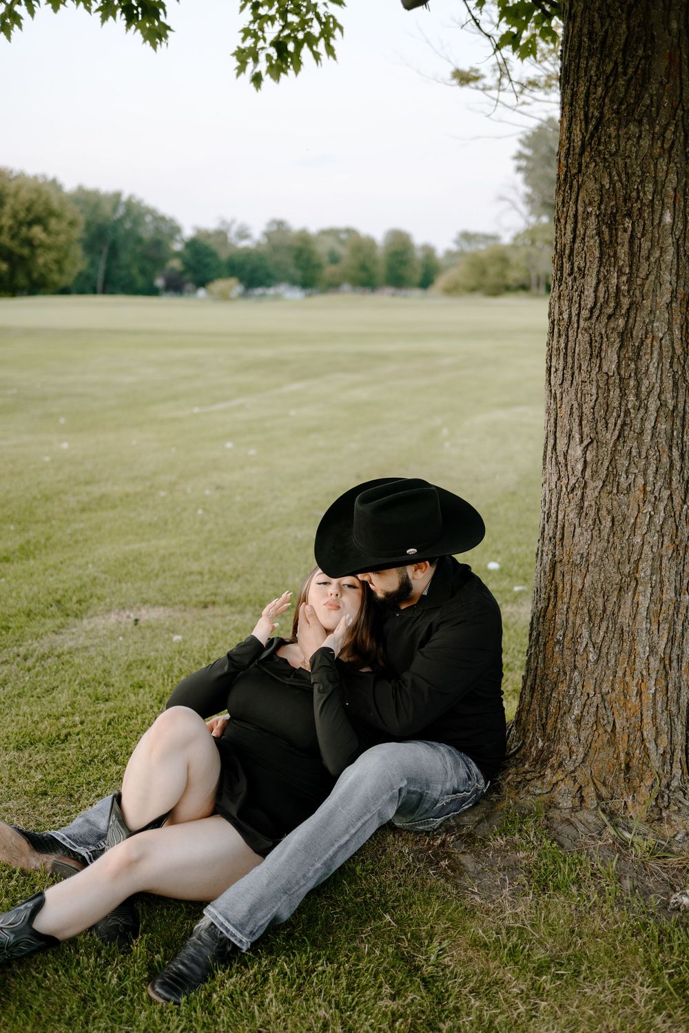 A couple rests intimately beneath a large tree in a peaceful park setting on a sunny day.