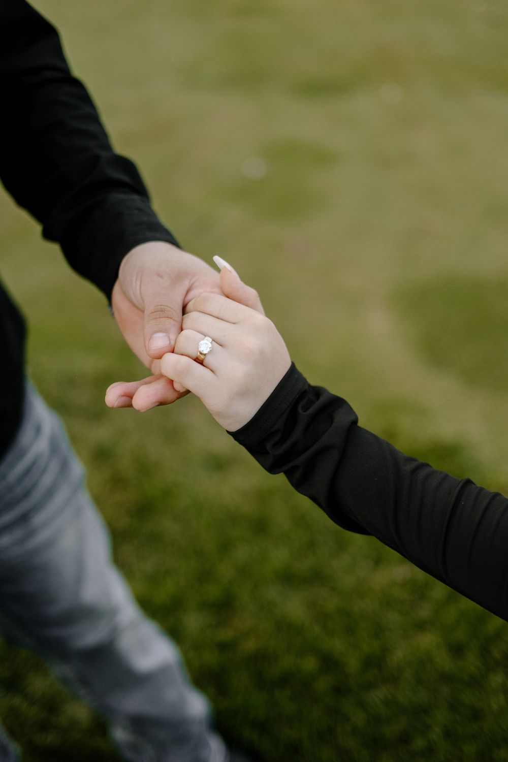 Close-up of clasped hands with an engagement ring against a green outdoor background.