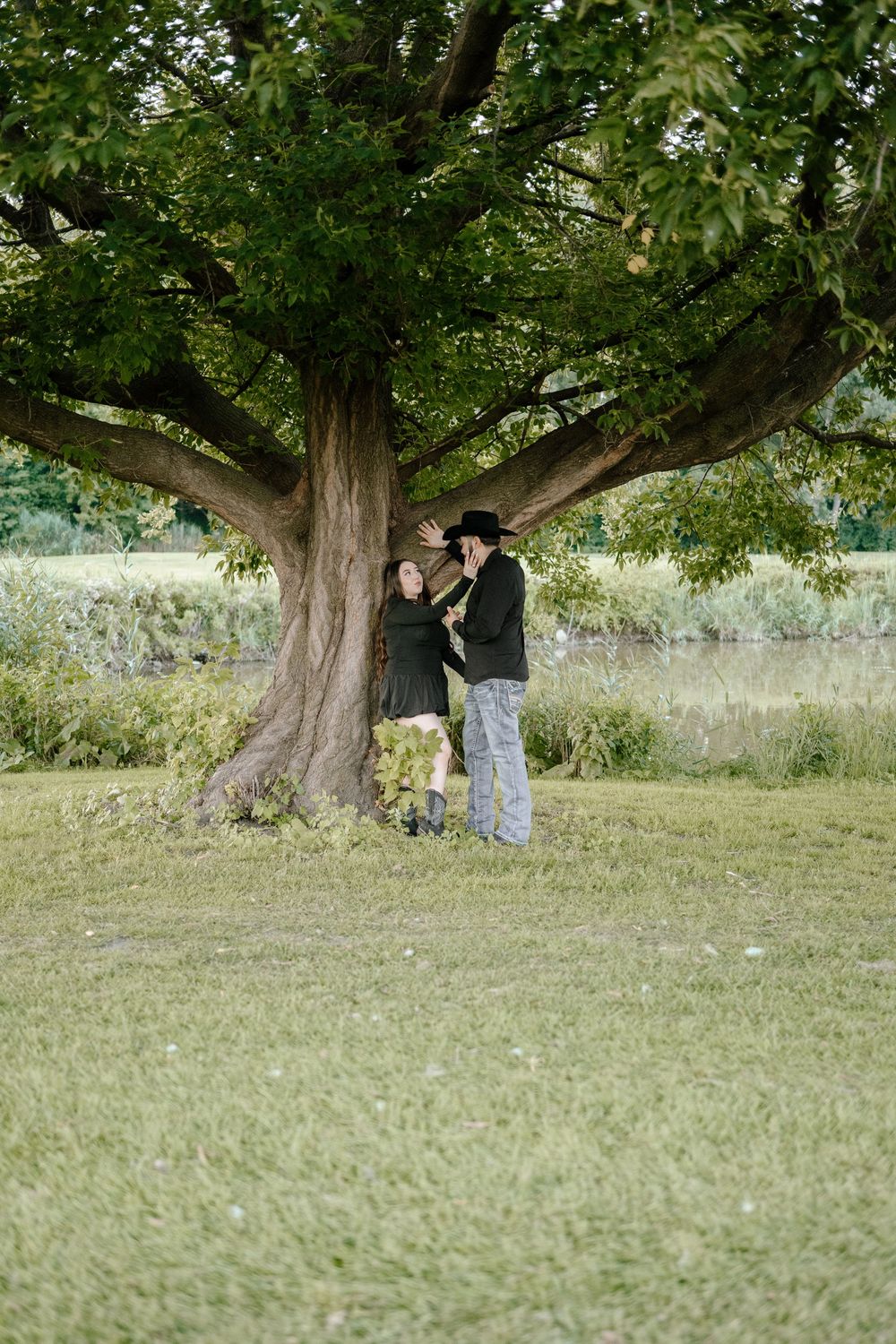 A couple stands together beneath a large sprawling tree in a grassy field at sunset.
