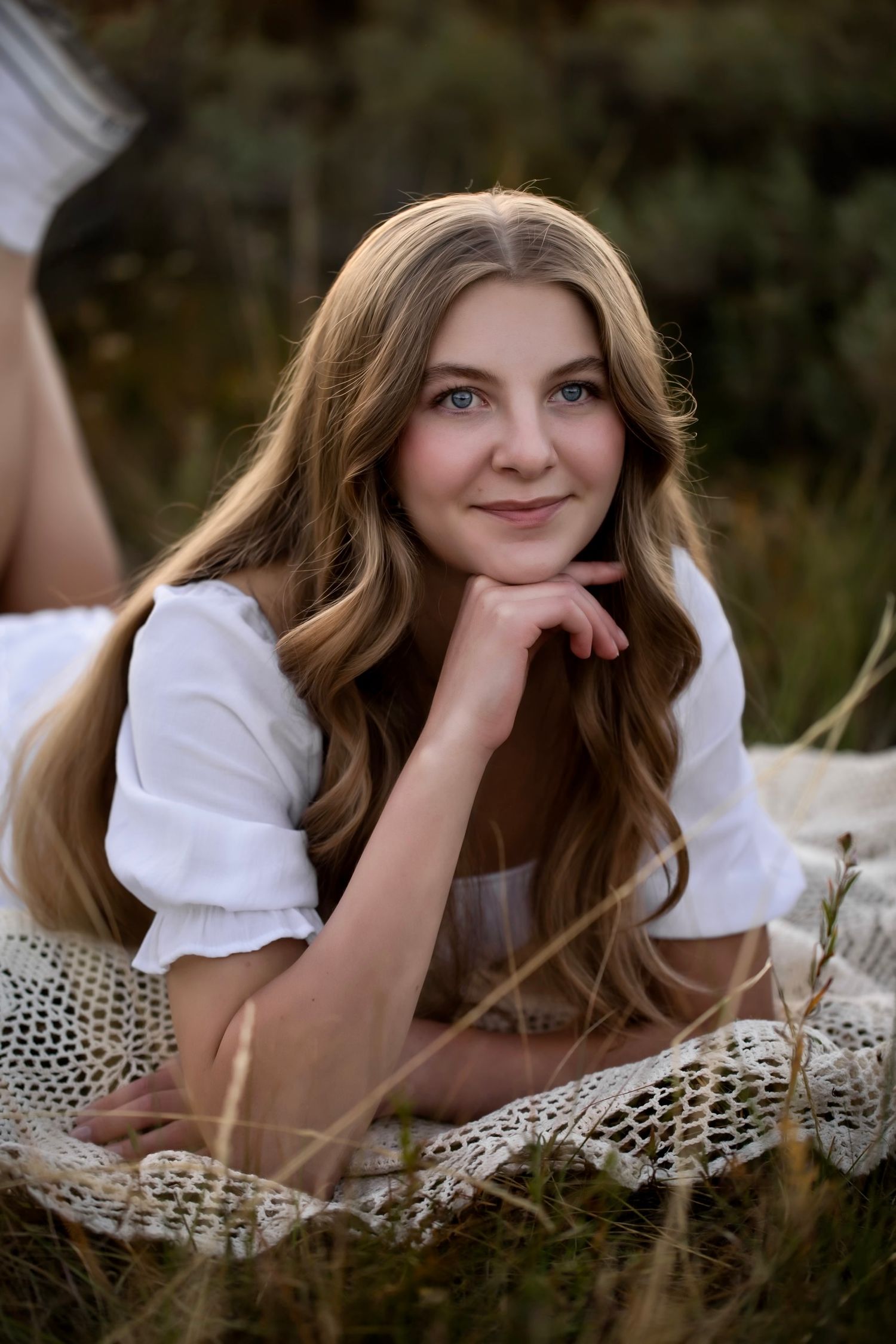 Young woman with long wavy hair lying on a blanket outdoors, smiling softly in a white top.