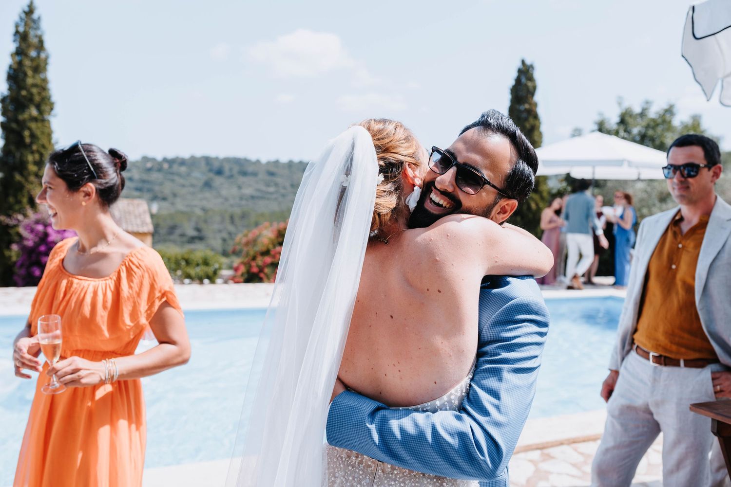 Hochzeitsgesellschaft in orangefarbener und blauer Kleidung gratuliert freudig der Braut am Pool der Finca Xiclati auf Mallorca.