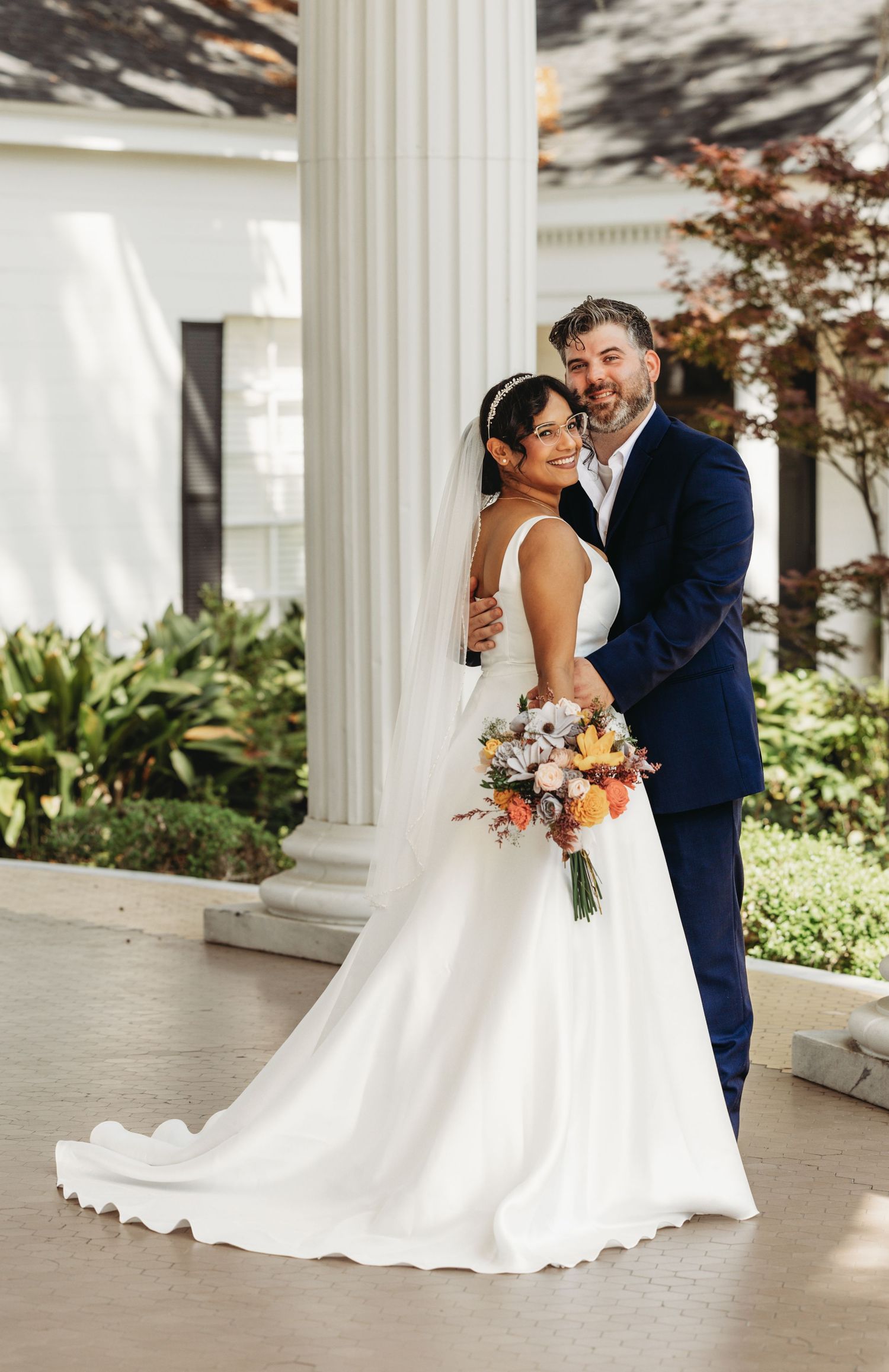 Newlyweds share intimate moments on a white columned porch with lush greenery.