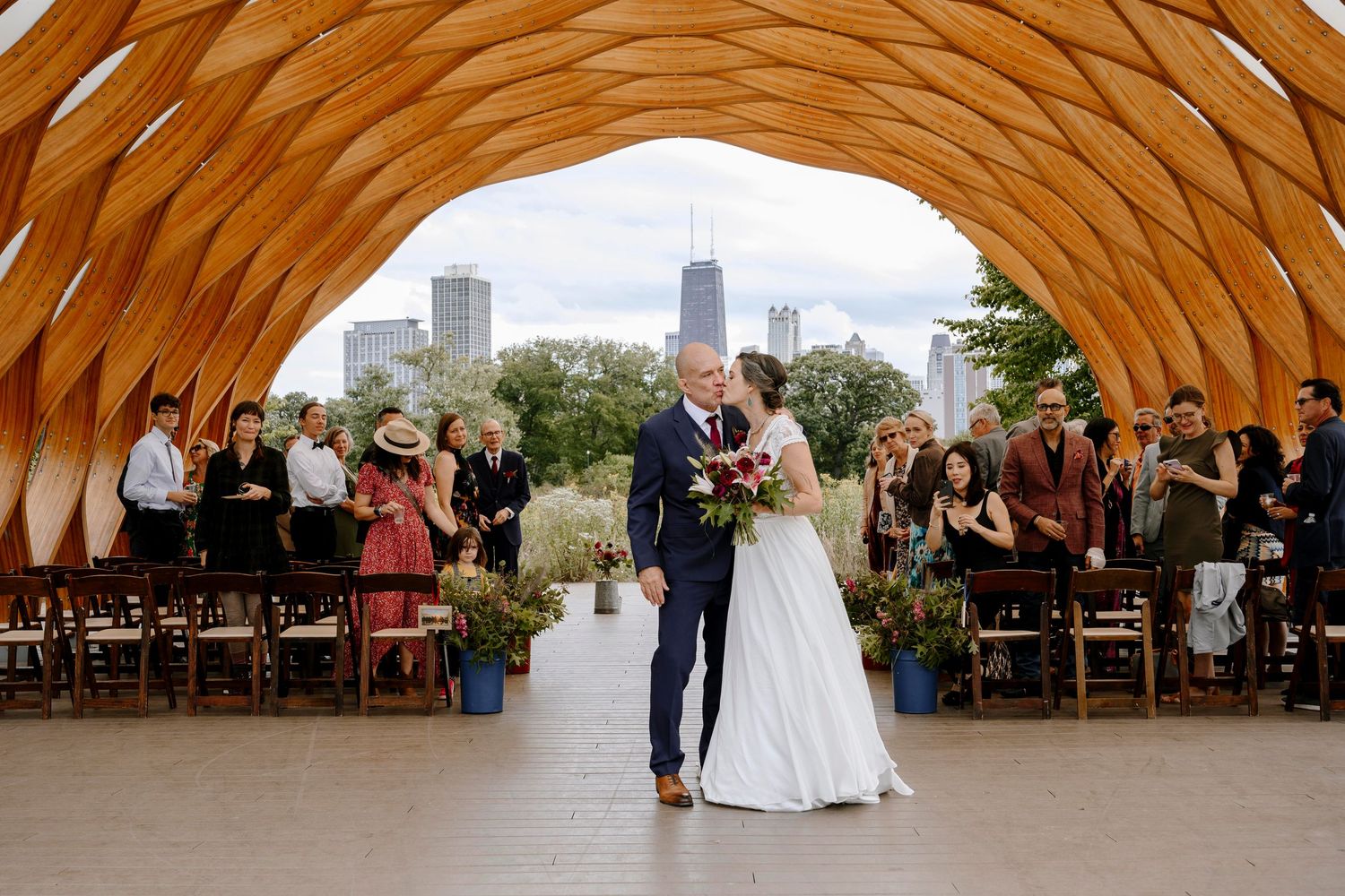 A couple sharing their first dance under a wooden dome structure with the Chicago skyline and wedding guests in the background.