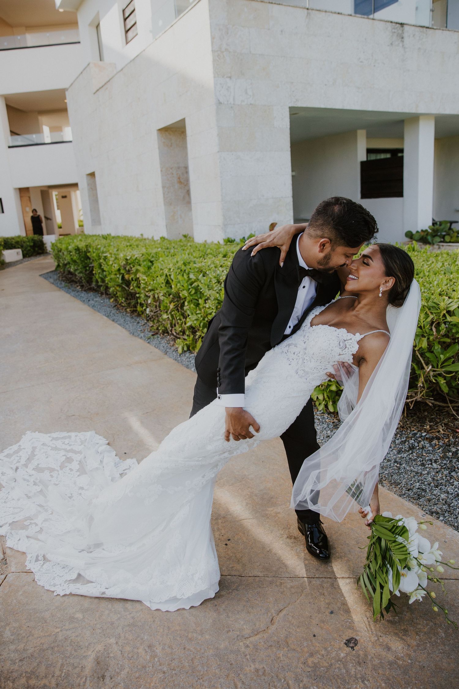 A romantic dip pose of newlyweds in formal attire against a white building backdrop.