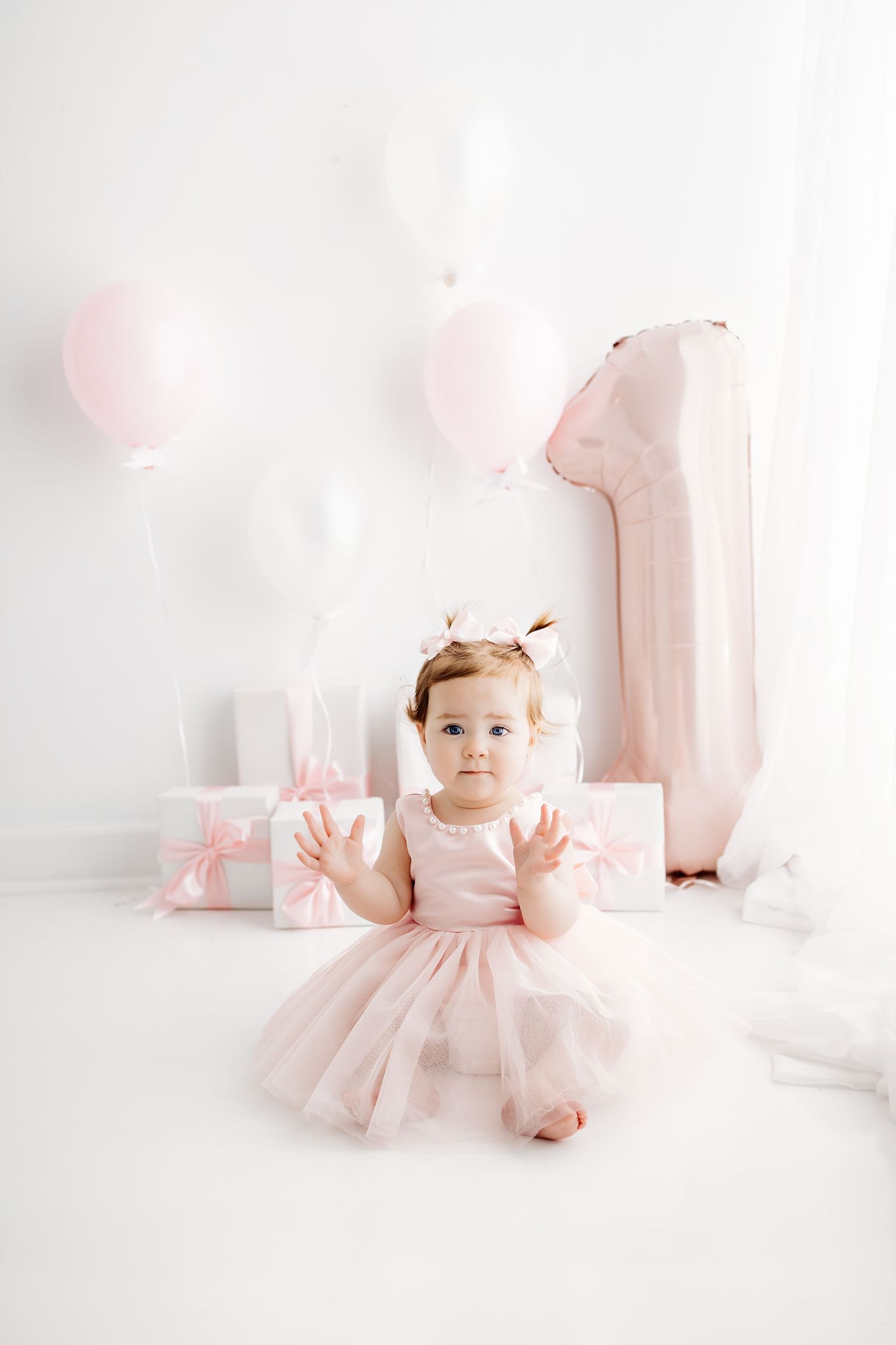 A toddler in a pink tulle dress and bow reaches up while standing in a light-filled room.