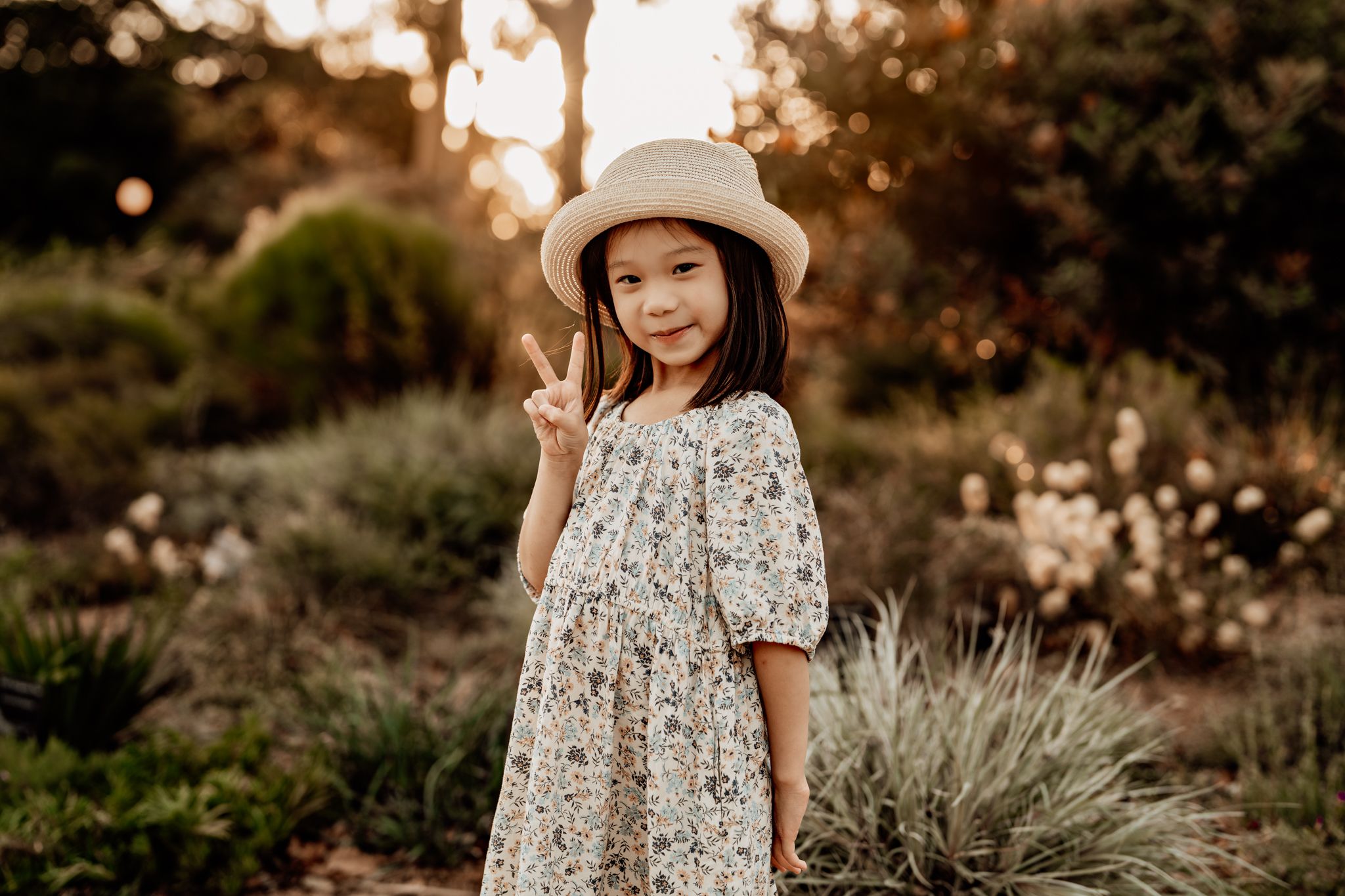 Young girl in floral dress poses in golden sunlit garden during sunset portrait session.