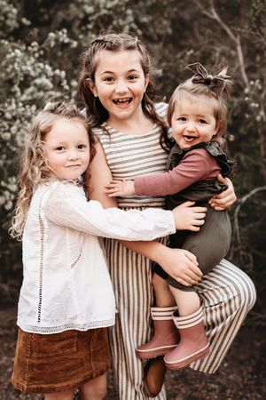 Three smiling children hugging outdoors in earth-toned clothing with white and striped patterns against a blurred background.