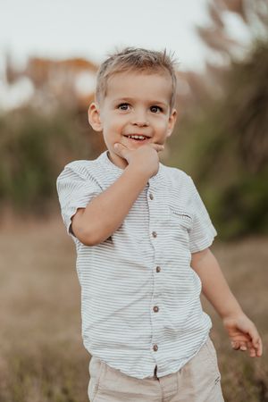 Young child in striped button-down shirt smiles while holding hand near face in outdoor portrait setting.