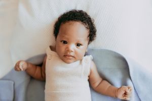 Baby wearing white sleeveless onesie lies on soft gray cushion looking at camera.