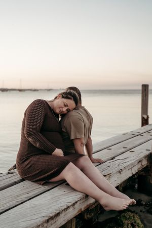 A person in brown sweater lays curled up on wooden dock by tranquil ocean at sunset.