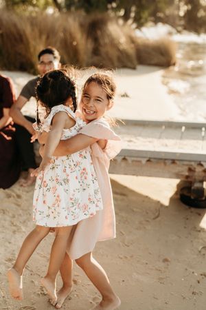Young children in floral dresses are laughing and playing together on a sunlit beach.